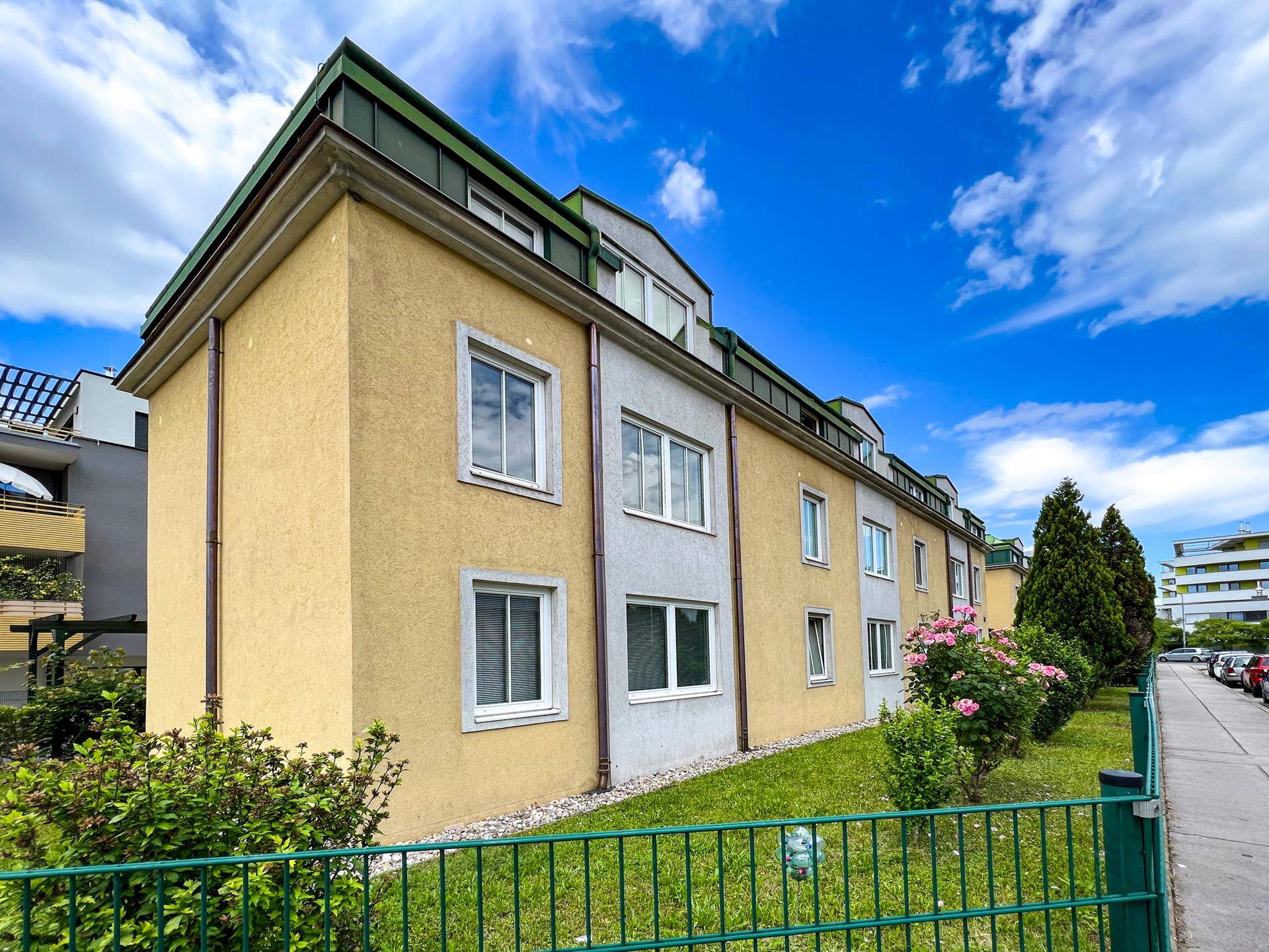 Seitenansicht des gelben Wohngebäudes mit gepflegter Grünfläche und blühenden Sträuchern.