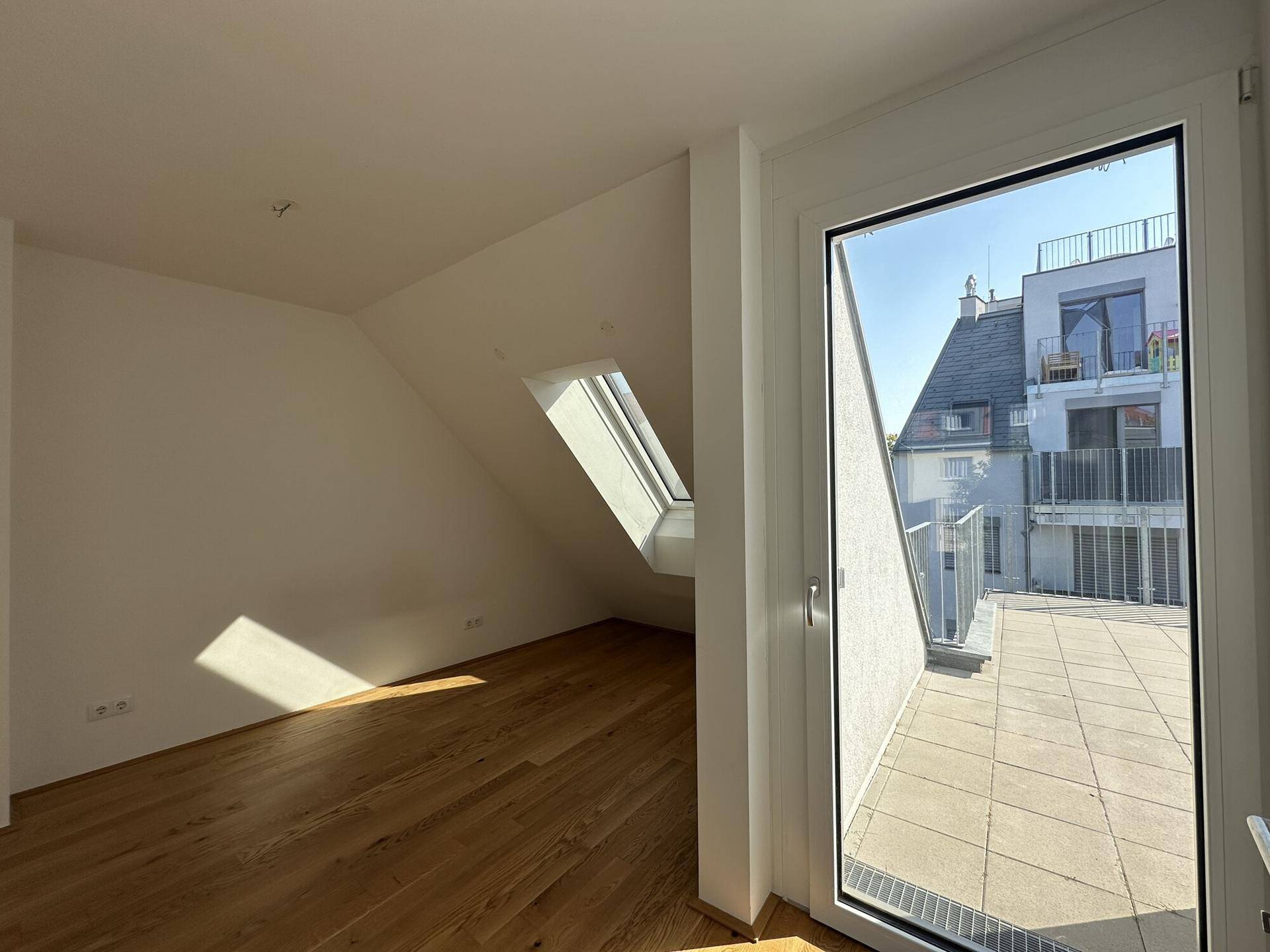 Helles Zimmer mit Dachschräge, Holzboden und Zugang zu einer Terrasse.