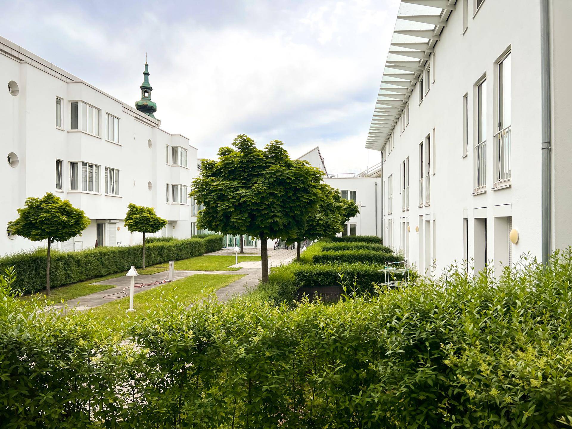 Grüner Innenhof mit Bäumen und Hecken, umgeben von modernen Wohngebäuden unter bewölktem Himmel.