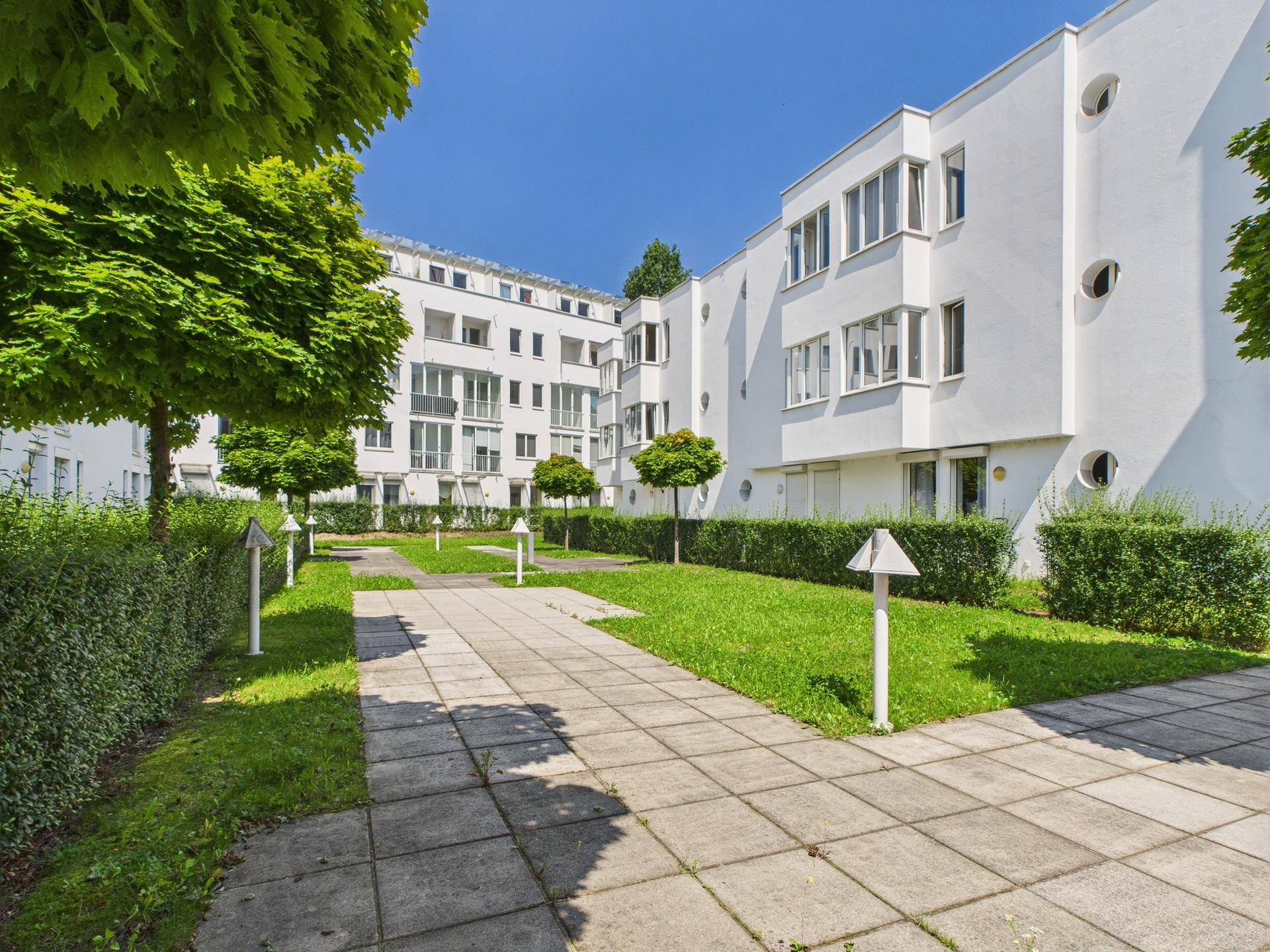 Sonniger Innenhof mit Rasenflächen, Bäumen und gepflasterten Wegen zwischen den Wohngebäuden.