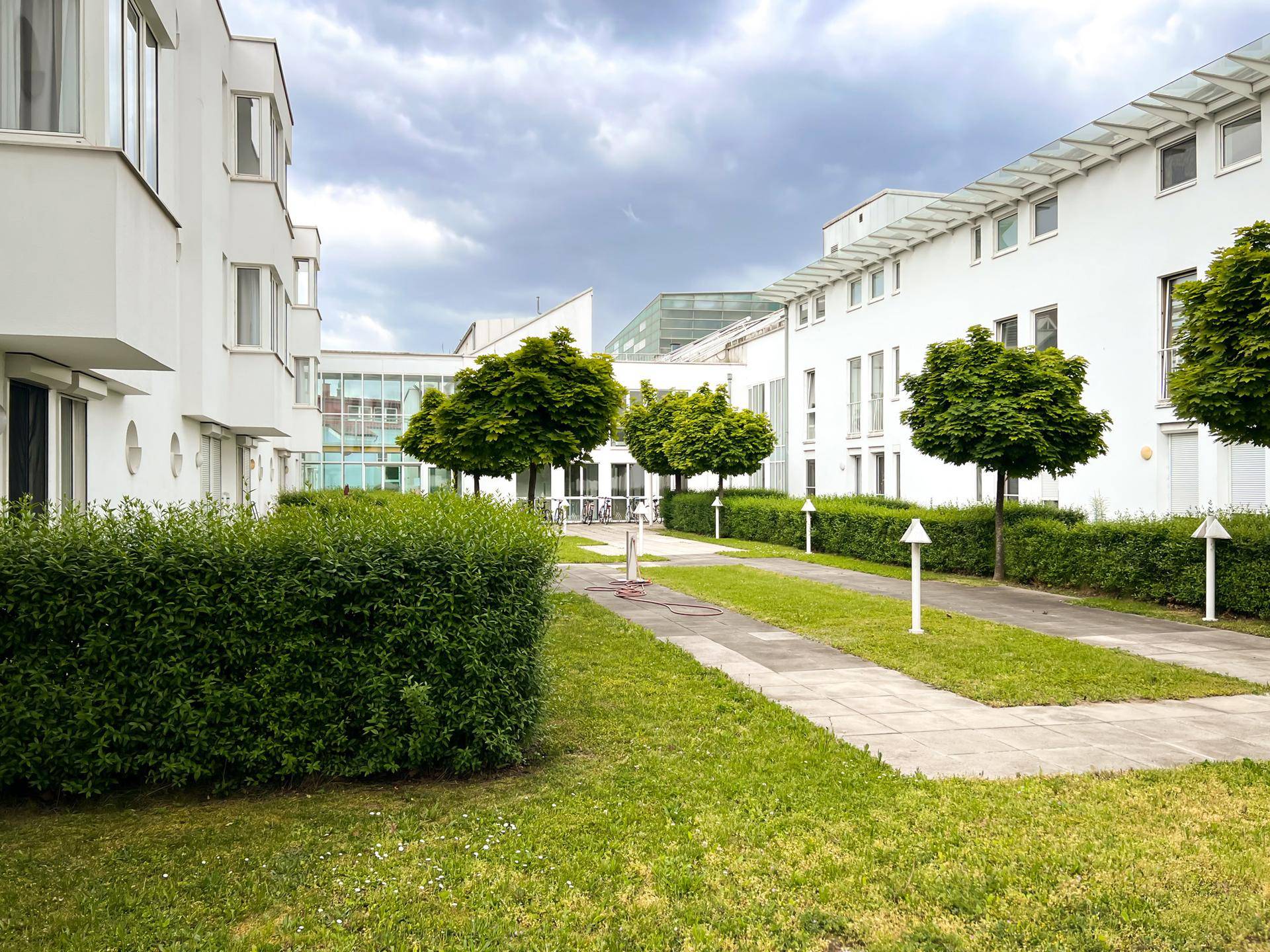 Grüner Innenhof mit Bäumen und Hecken, umgeben von modernen Wohngebäuden unter bewölktem Himmel.