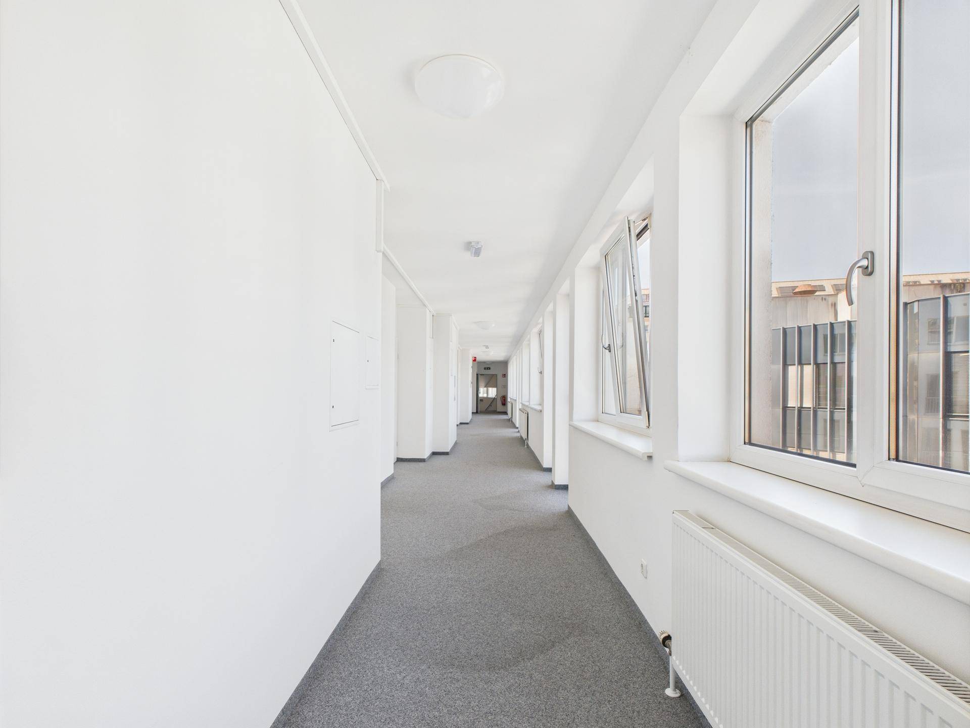 Langer, heller Flur mit Fenstern und grauem Teppichboden in einem modernen Gebäude.