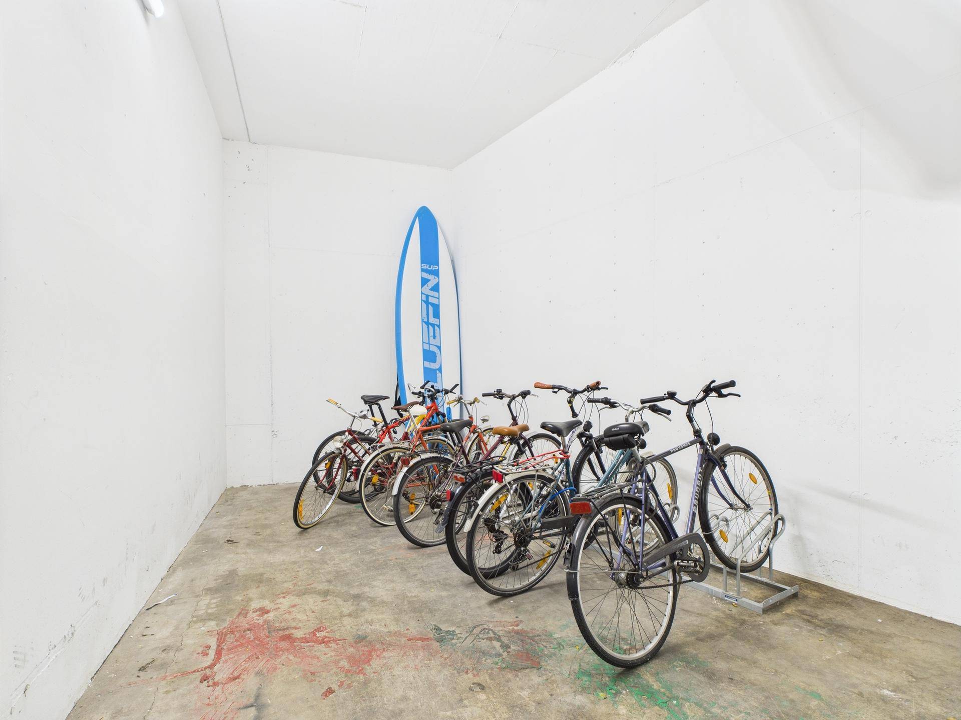 Heller Fahrradabstellraum mit mehreren Fahrrädern und einem Surfbrett an der Wand.