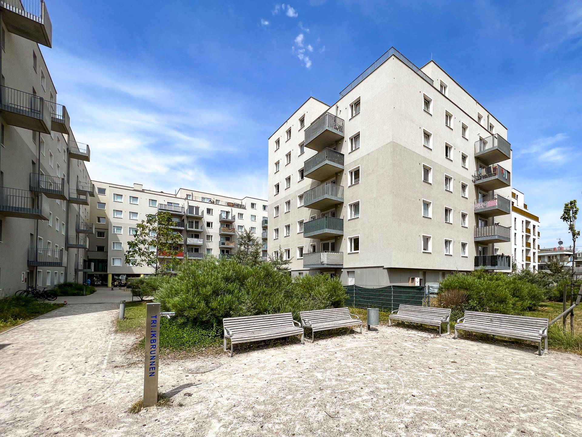 Ruhiger Innenhof mit Sitzbänken und einem sandigen Weg, umgeben von modernen Wohngebäuden.