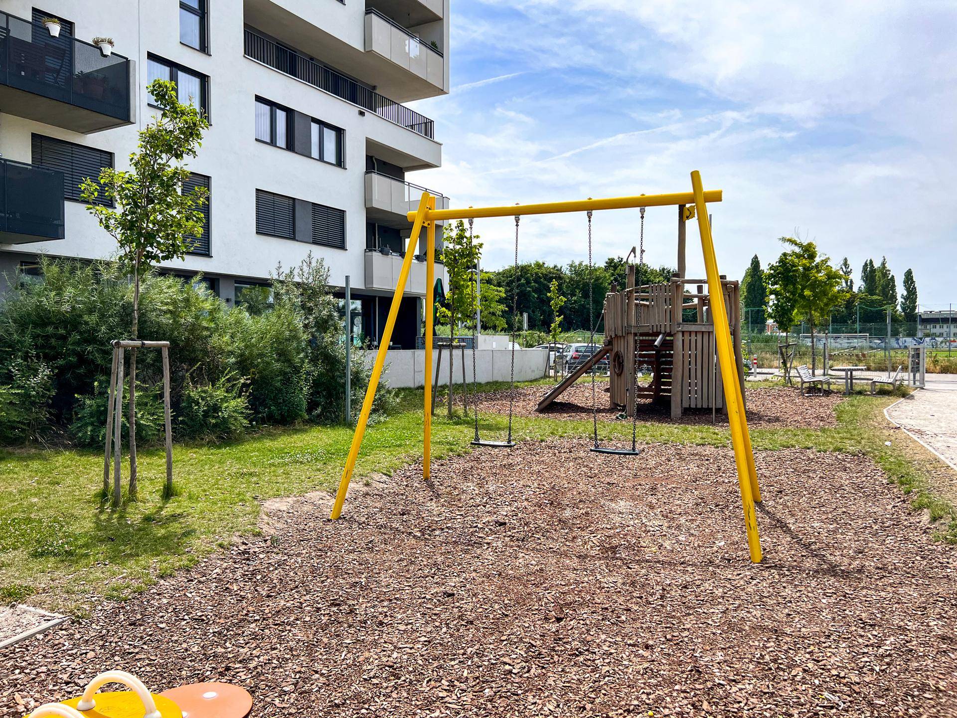 Kinderspielplatz mit gelber Schaukel und Holzspielgeräten, umgeben von Grünflächen und Wohngebäuden.