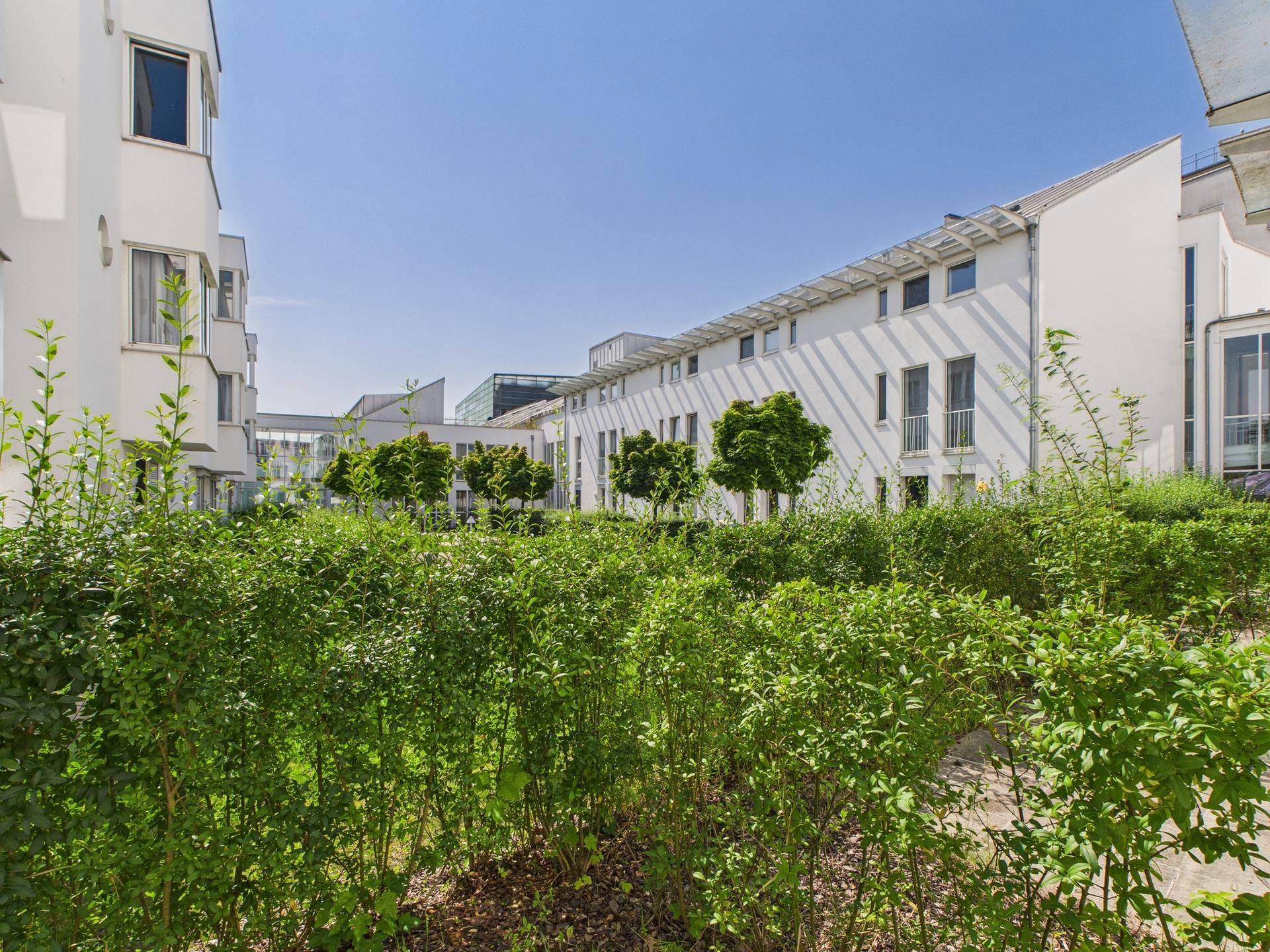 Gepflegter Gartenbereich mit grünen Hecken und Bäumen zwischen modernen Wohngebäuden unter blauem Himmel.