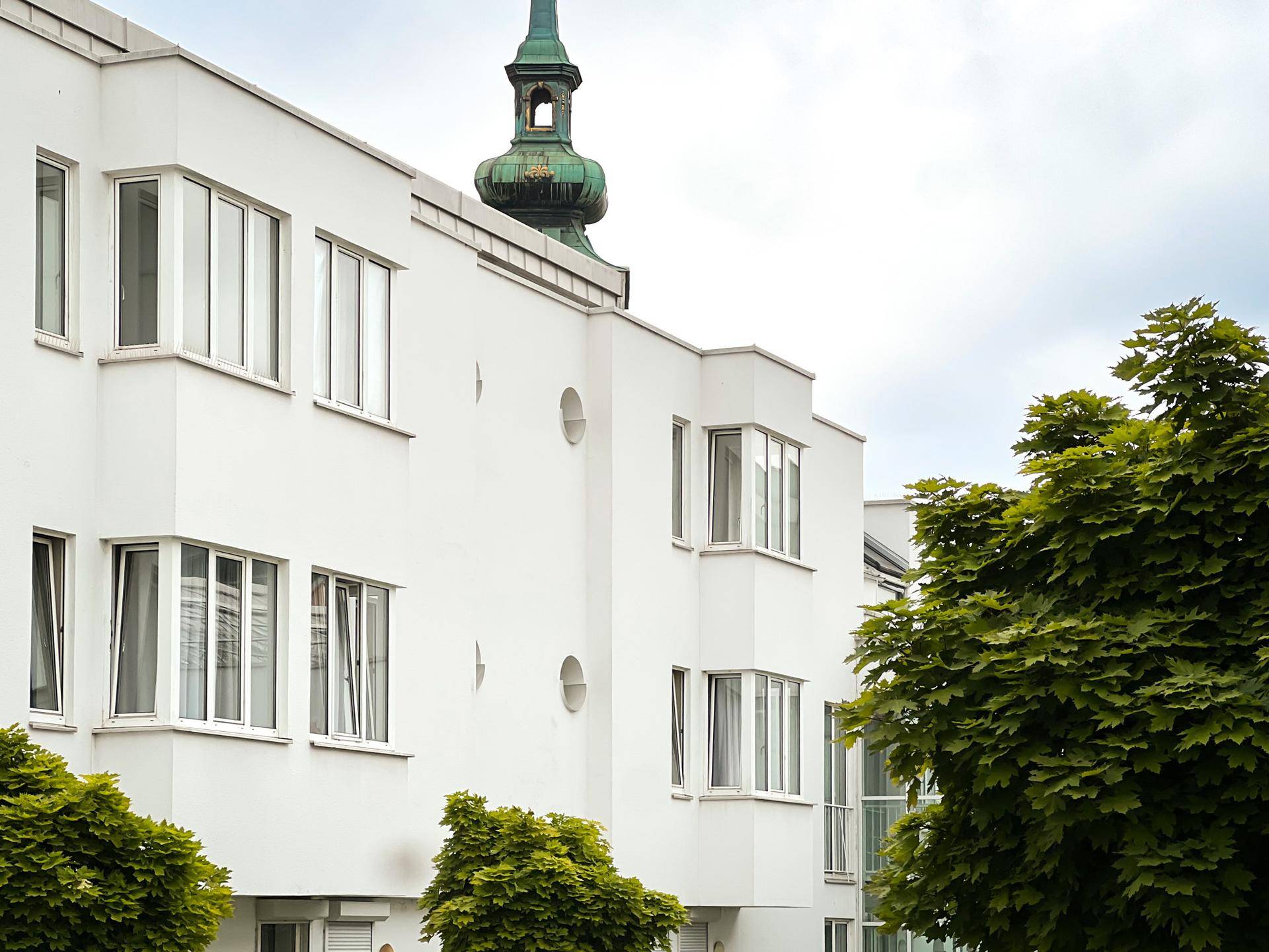 Moderne weiße Gebäudefassade mit Erkerfenstern und einem Kirchturm im Hintergrund unter bewölktem Himmel.