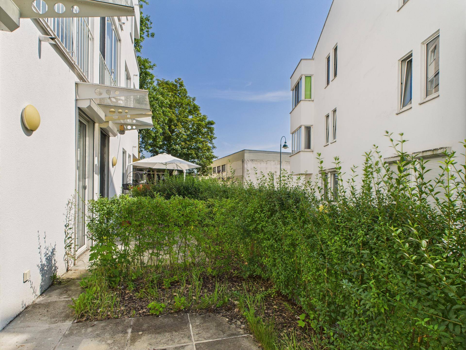 Außenbereich mit gepflasterter Terrasse und grünen Hecken, umgeben von modernen Gebäuden und einem Sonnenschirm.