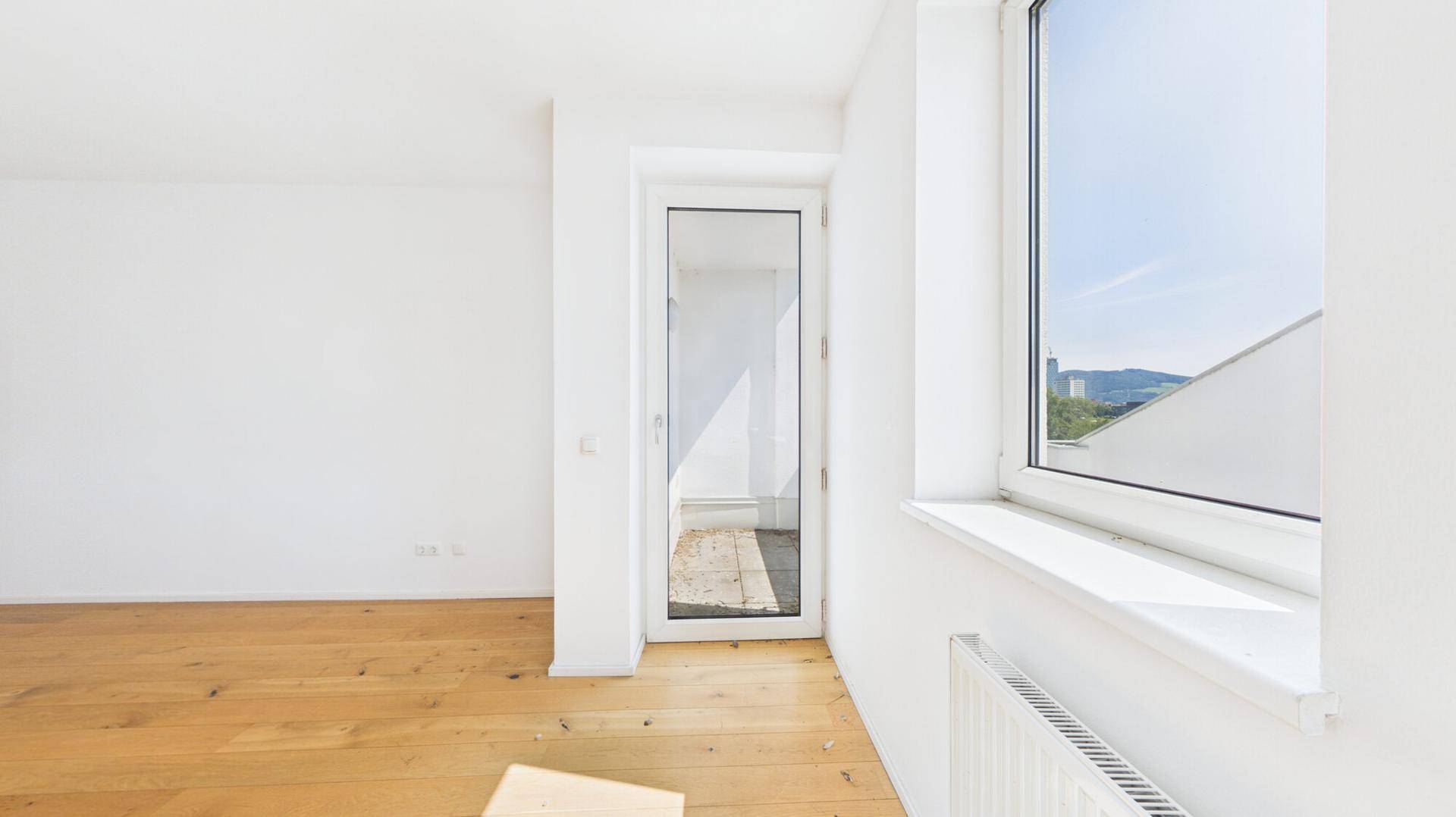 Helle Ecke eines Zimmers mit großem Fenster, Heizkörper und Zugang zu einem Außenbereich.