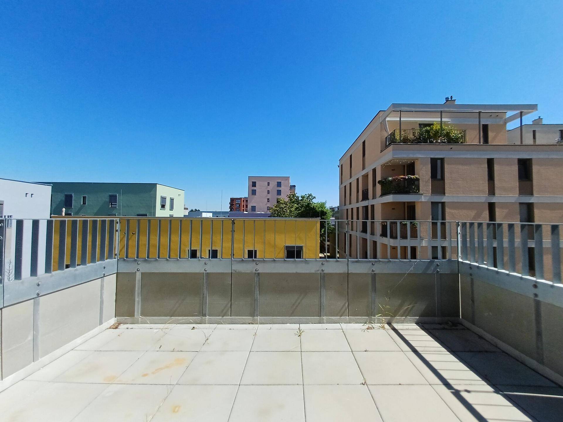 Geräumige Dachterrasse mit Fliesenboden und Blick auf moderne Wohngebäude unter blauem Himmel.