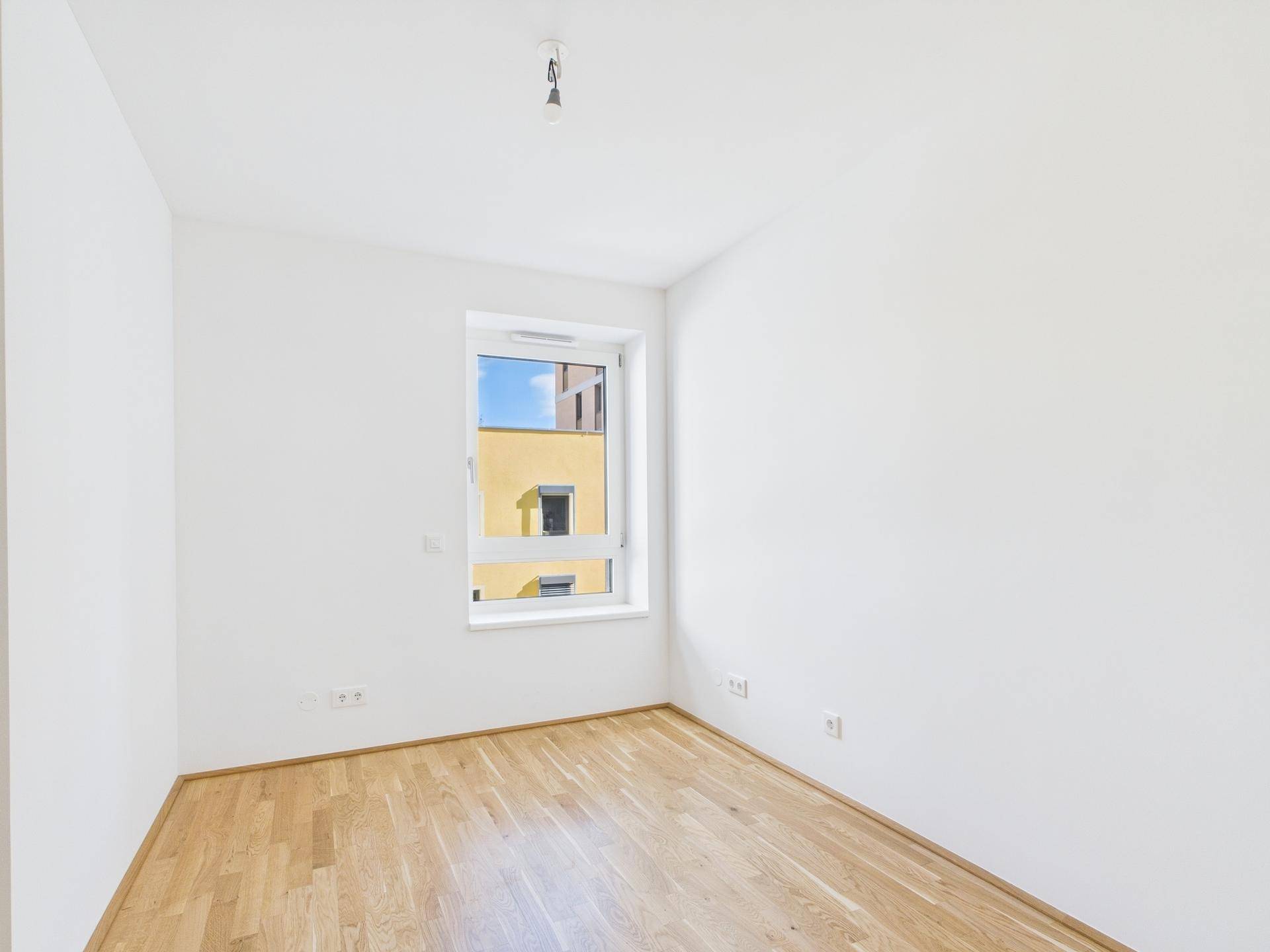 Helles, leeres Zimmer mit Parkettboden und einem Fenster, das den Blick auf ein gelbes Gebäude freigibt.