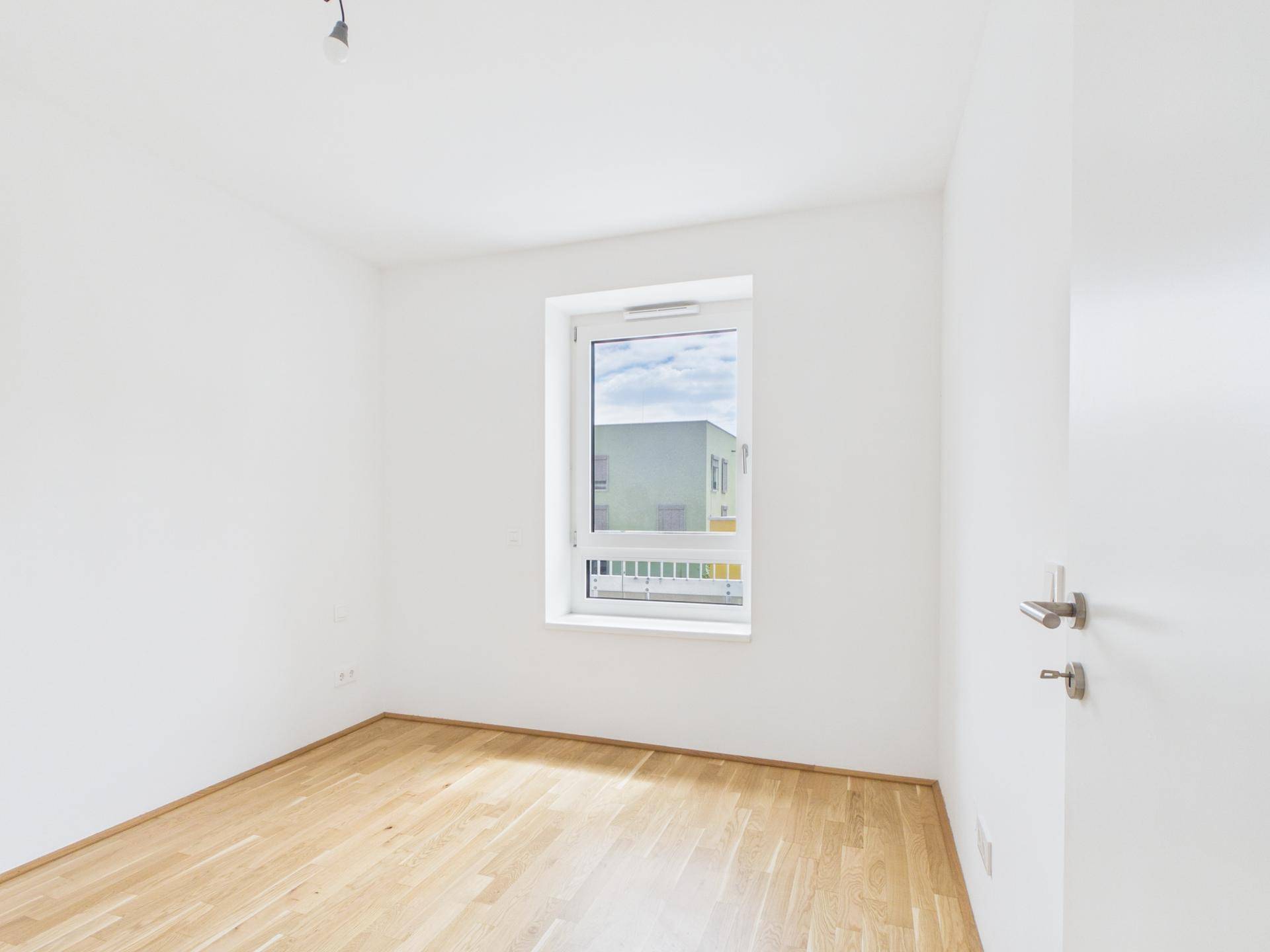 Leeres, helles Zimmer mit Parkettboden und einem Fenster, das viel Tageslicht hereinlässt.