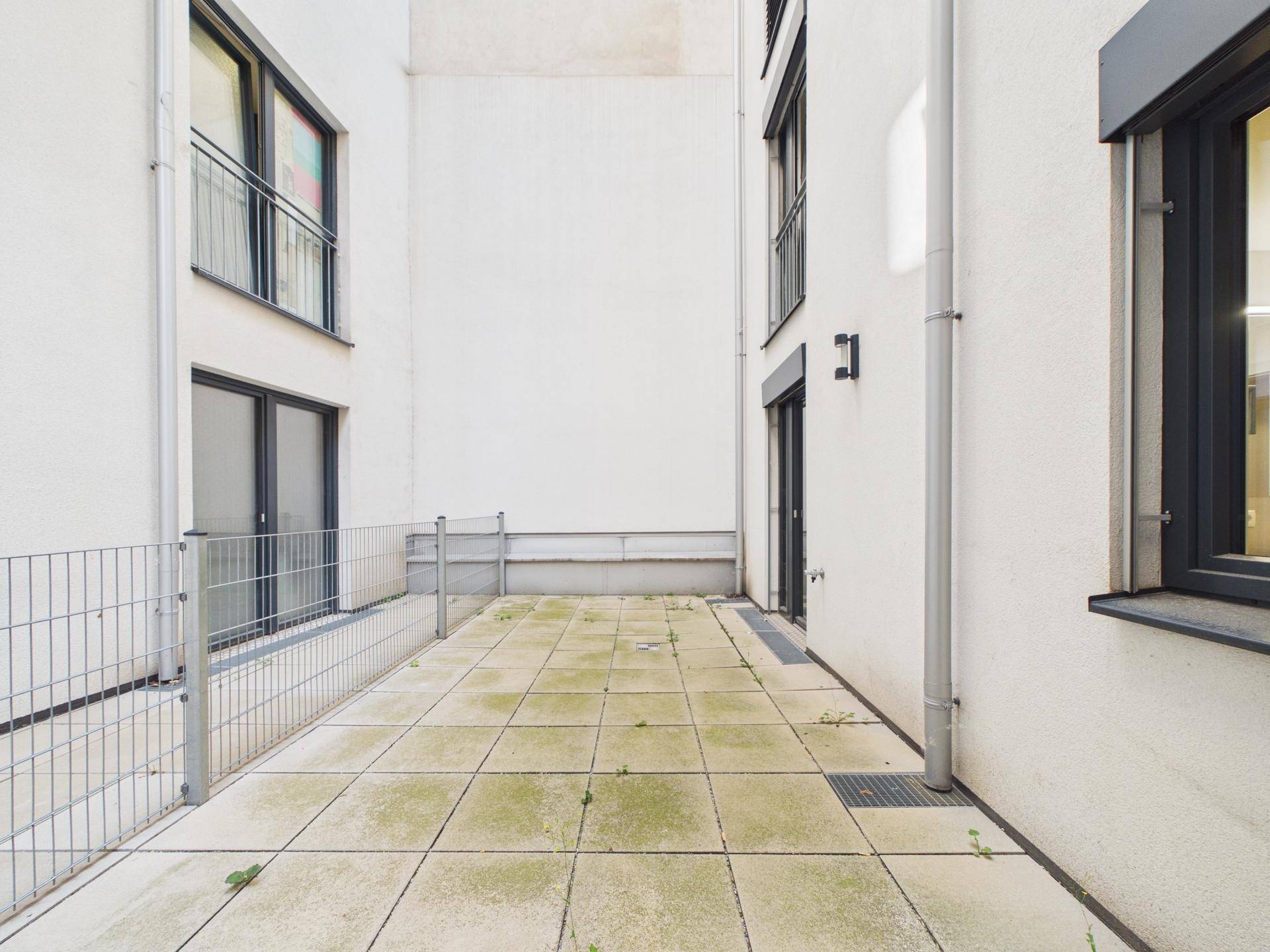 Lange, schmale Terrasse mit Fliesenboden und Metallzaun, umgeben von Gebäudefassaden.