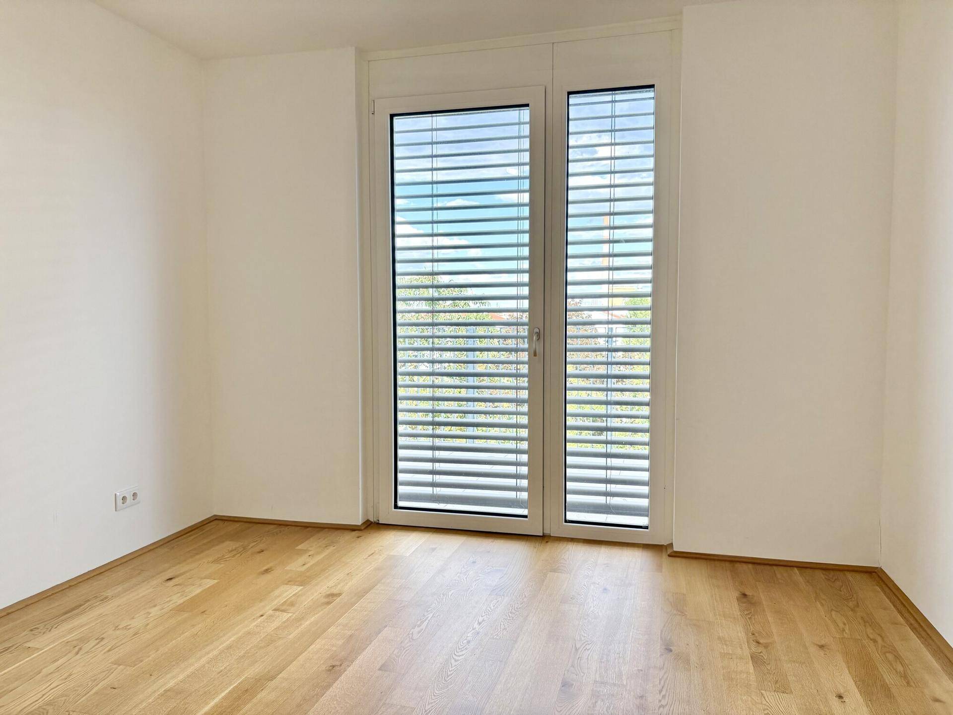 Helles Schlafzimmer mit Holzboden und großem Fenster mit Außenjalousien.
