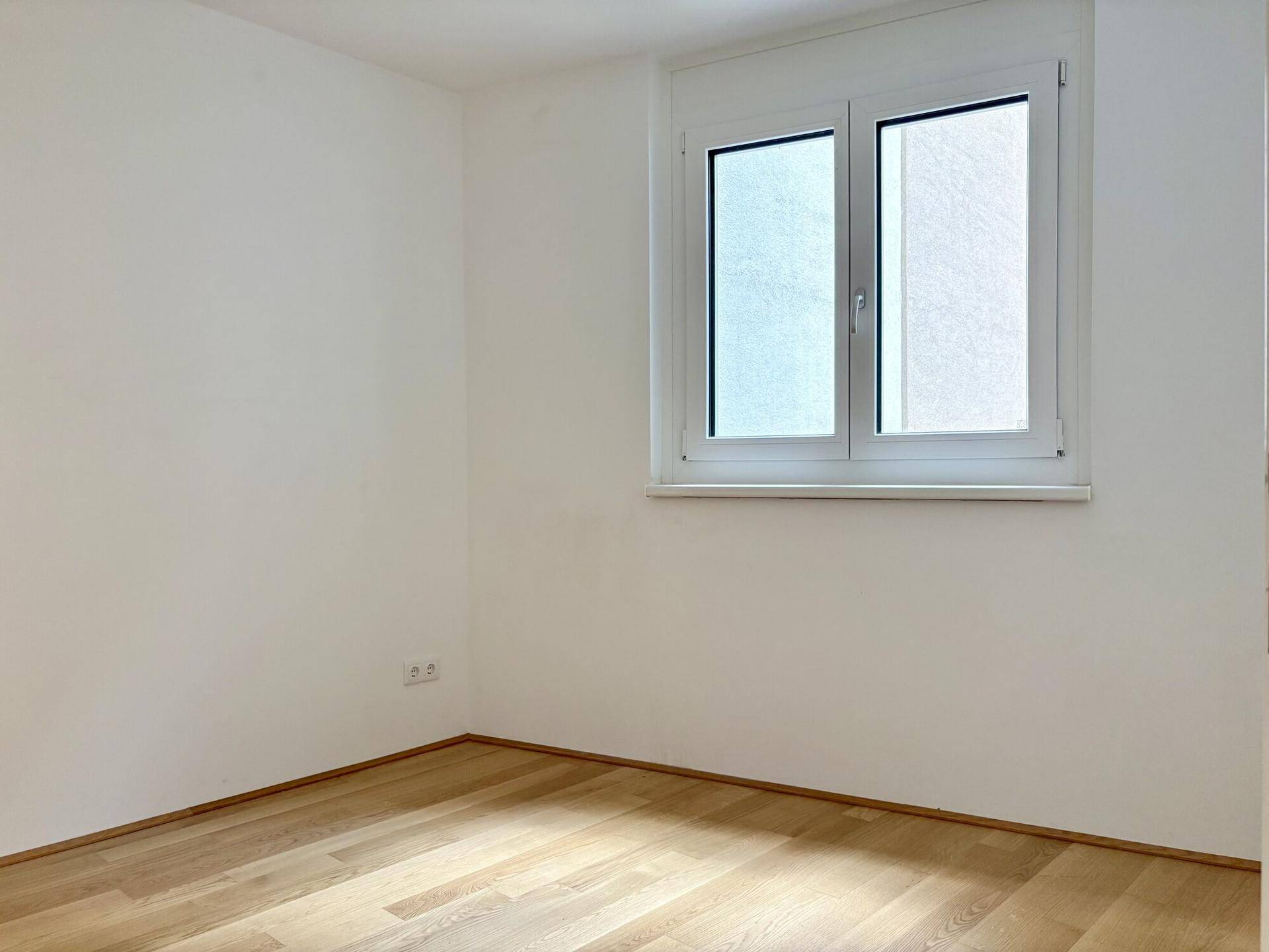 Ruhiges Schlafzimmer mit hellem Parkettboden und einem Fenster, das viel Tageslicht hereinlässt.