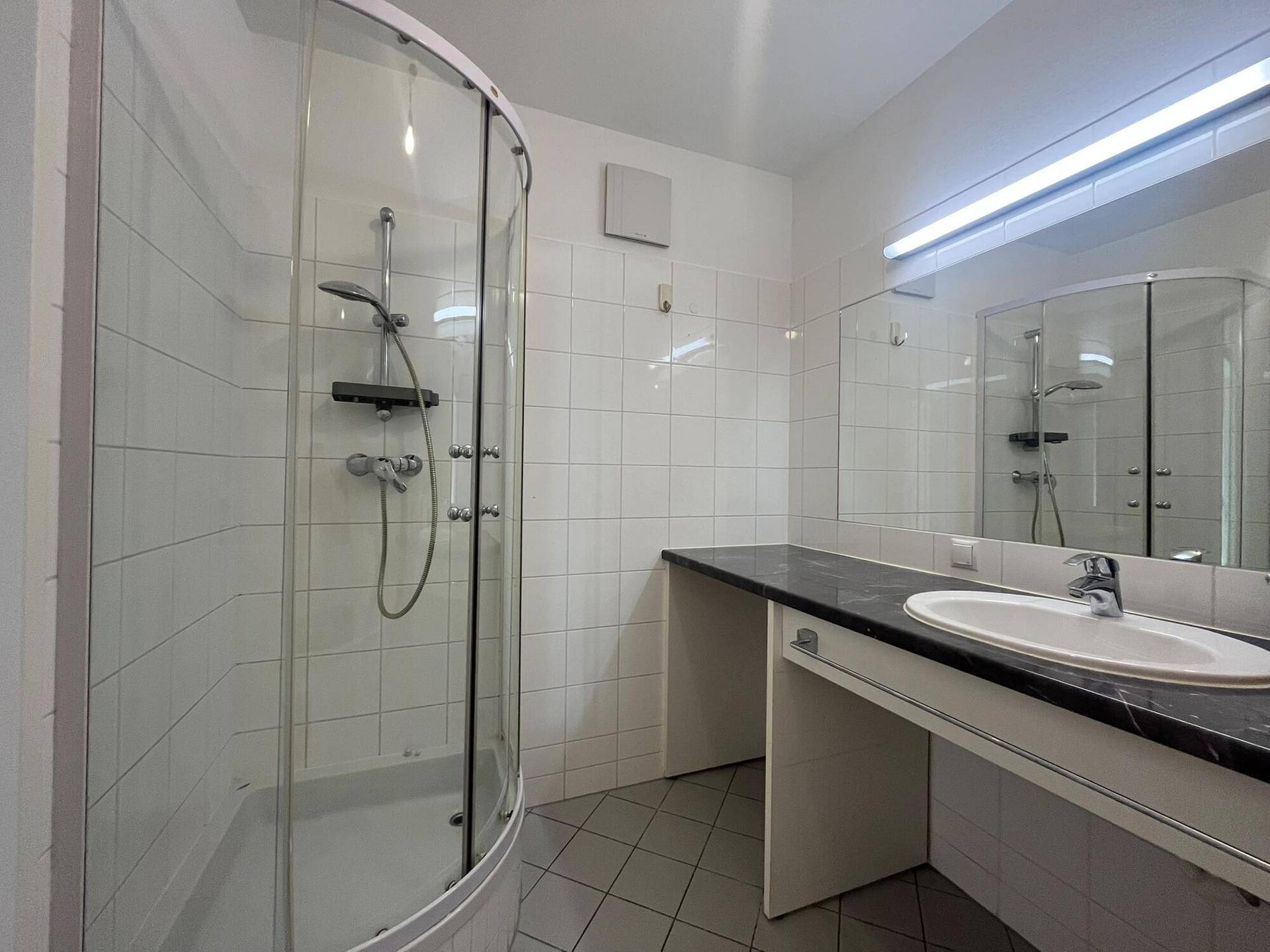 Badezimmer mit Eckdusche, Einzelwaschbecken und dunkler Arbeitsplatte, komplett gefliest.
