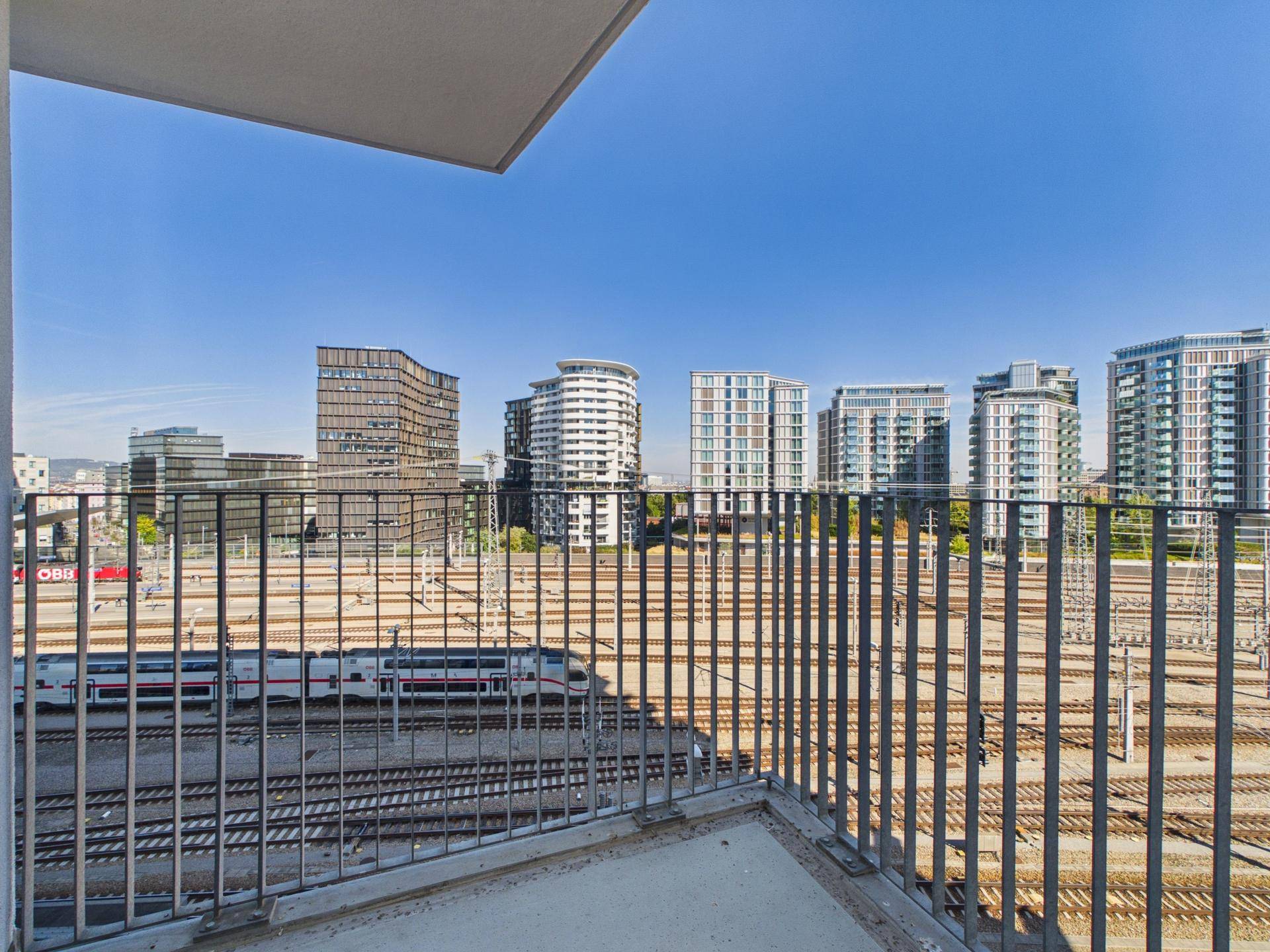 Balkon mit Blick auf die Stadtlandschaft, moderne Gebäude und Bahngleise unter blauem Himmel.