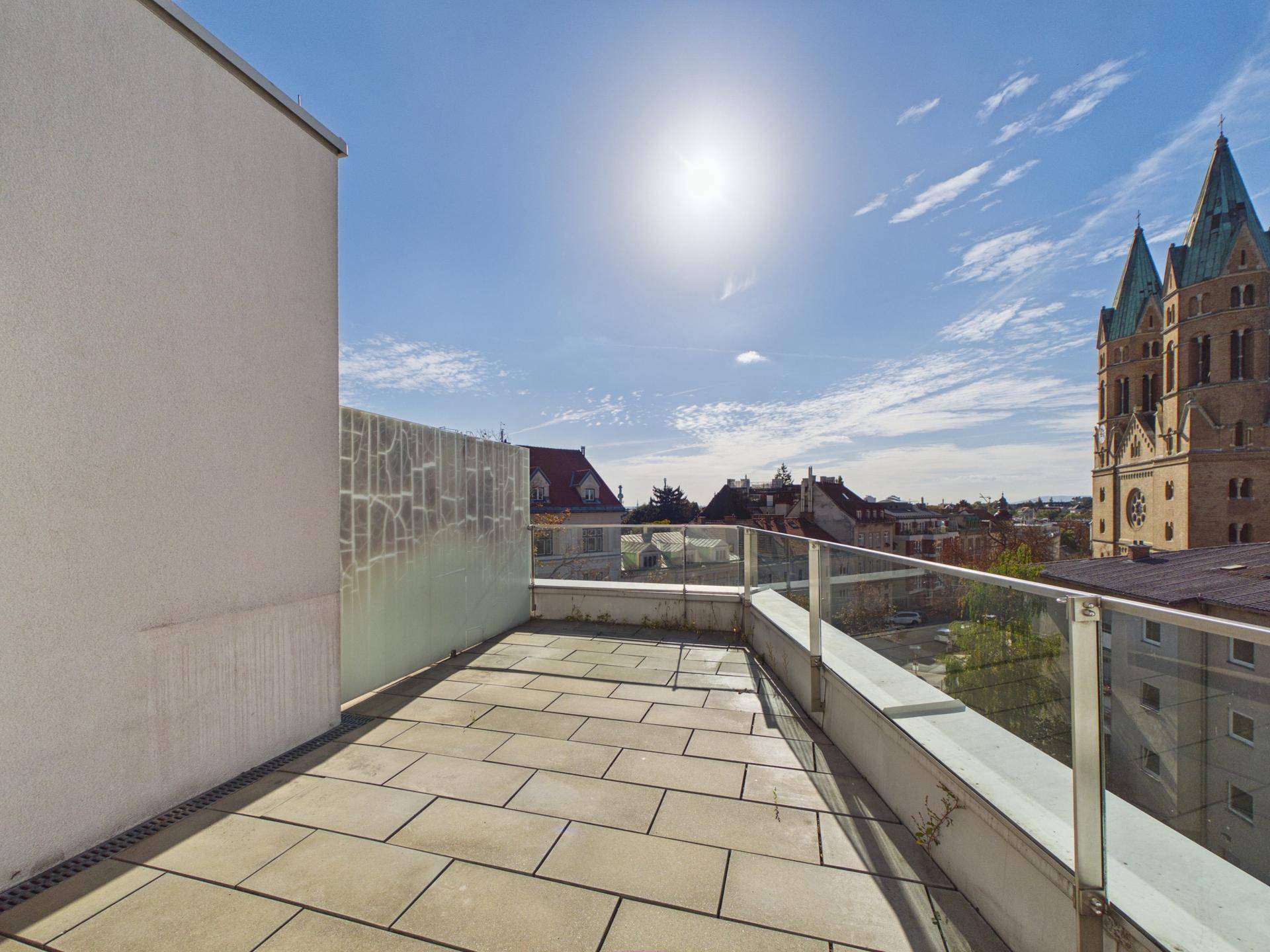 Große Dachterrasse mit Fliesenboden, Glasgeländer und weitem Blick über die Stadt und Kirche.