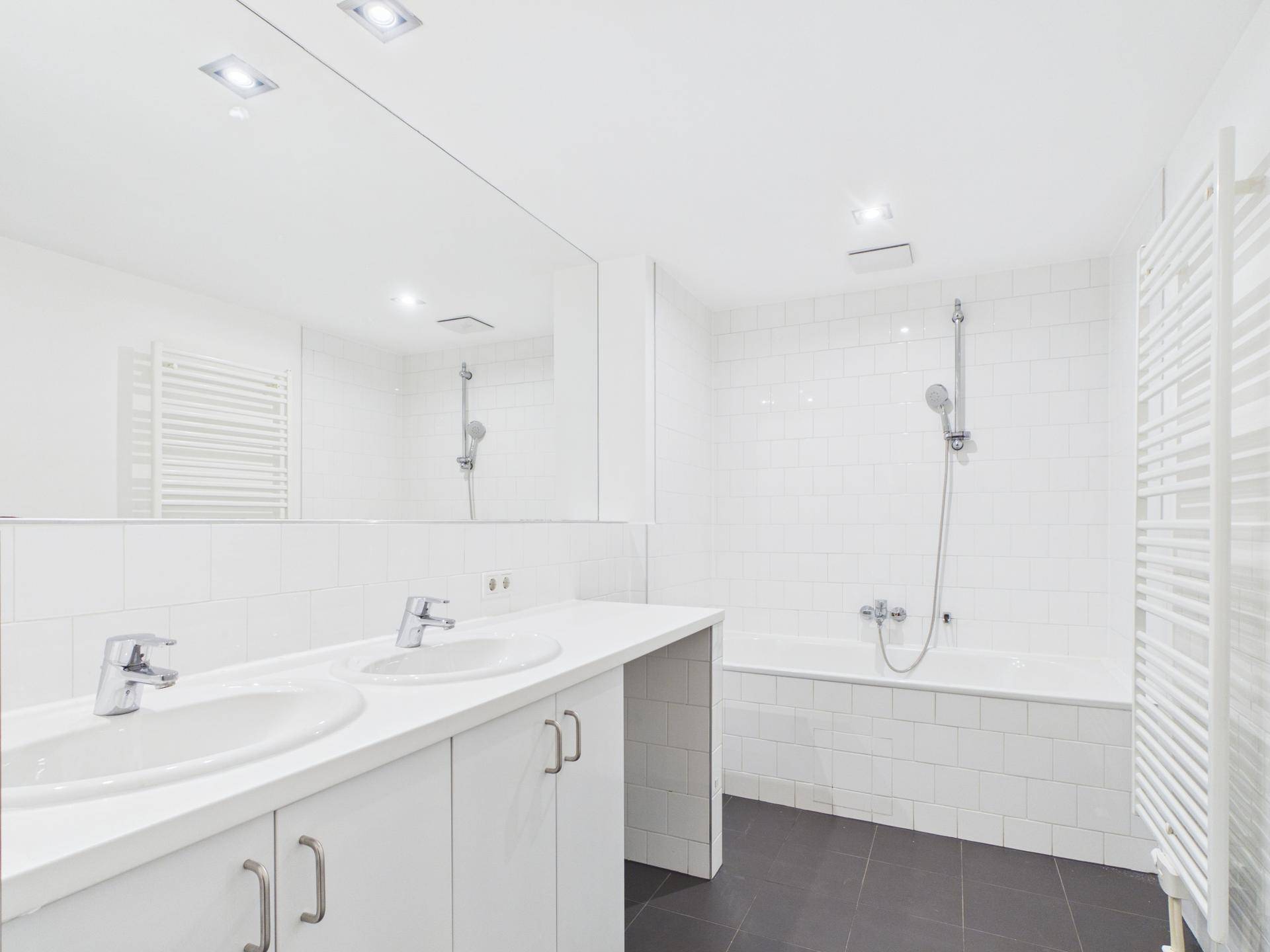 Modernes Badezimmer mit weißen Fliesen, Doppelwaschbecken, großer Spiegelfläche und Badewanne.