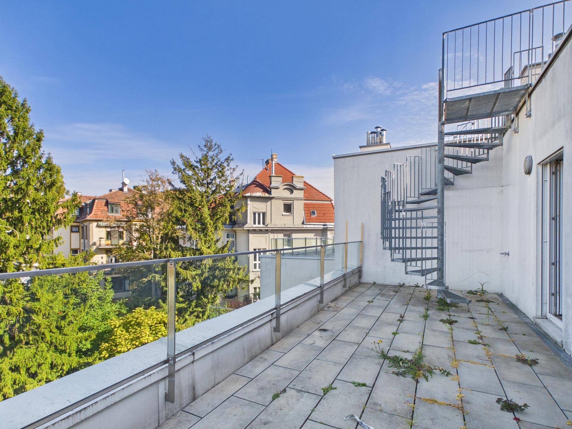Terrasse mit Fliesenboden, Glasgeländer und einer Wendeltreppe, umgeben von Bäumen und Häusern.