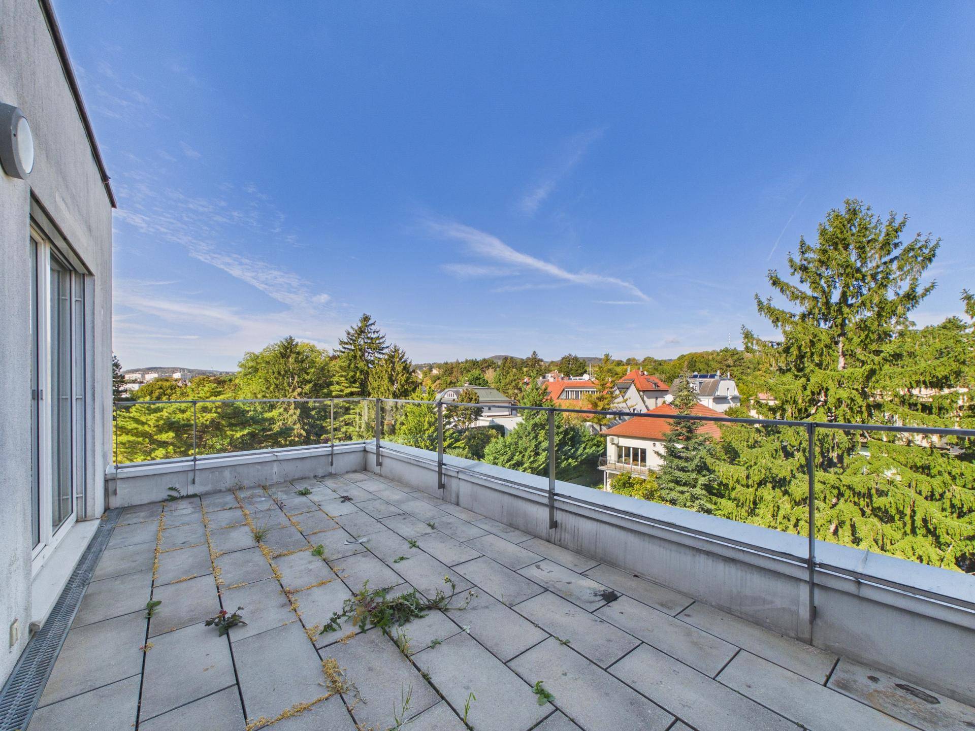 Geräumige Terrasse mit Fliesenboden und Glasgeländer, umgeben von viel Grün und Häusern.