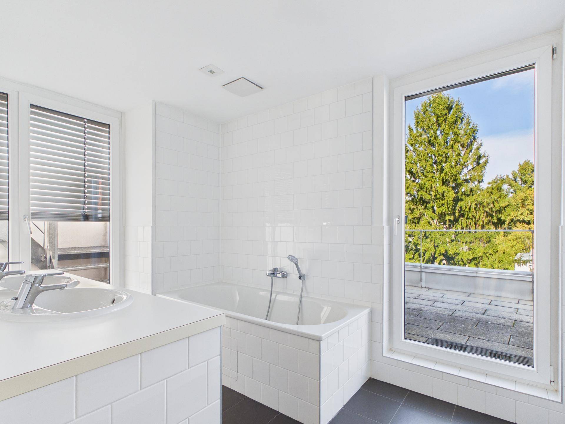 Helles Badezimmer mit Badewanne, Waschtisch und Fenster mit Blick ins Grüne.