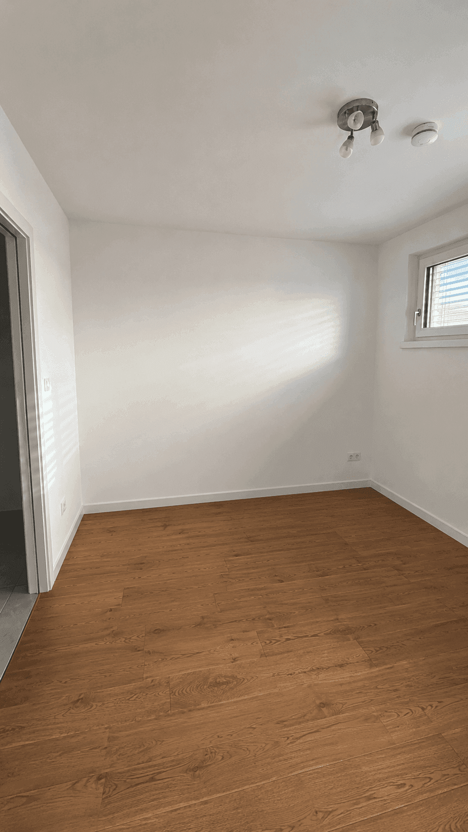 Helles Schlafzimmer mit Holzboden und einem Fenster mit Jalousien, bereit zur Einrichtung.