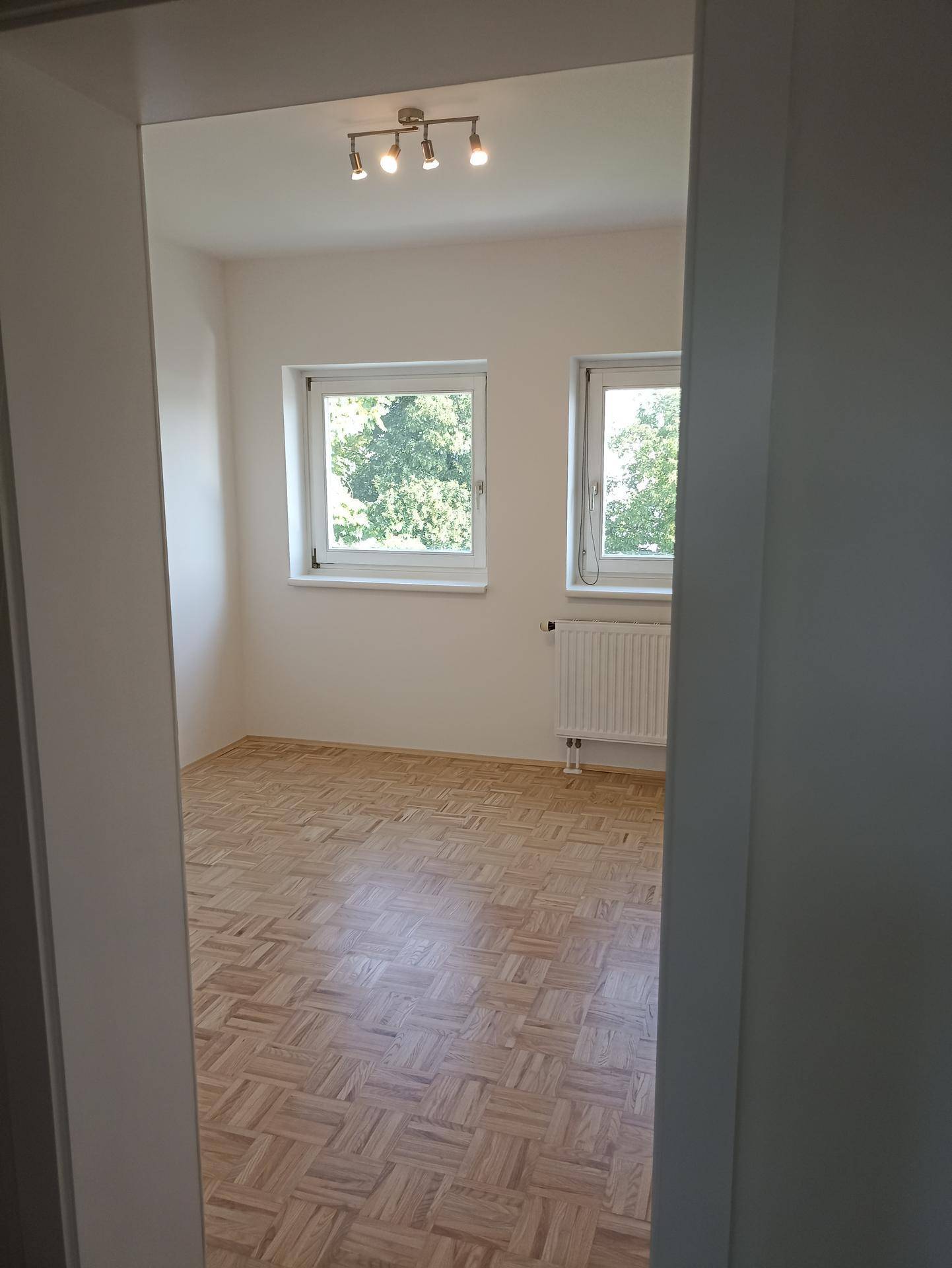 Helles Zimmer mit Parkettboden und zwei Fenstern, die einen Blick ins Grüne bieten.