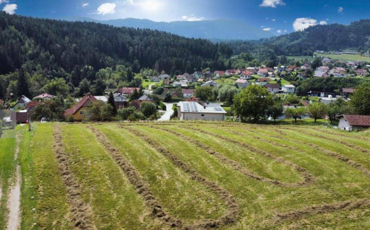 Weitläufiges gemähtes Feld mit Blick auf eine ländliche Siedlung und bewaldete Hügel.