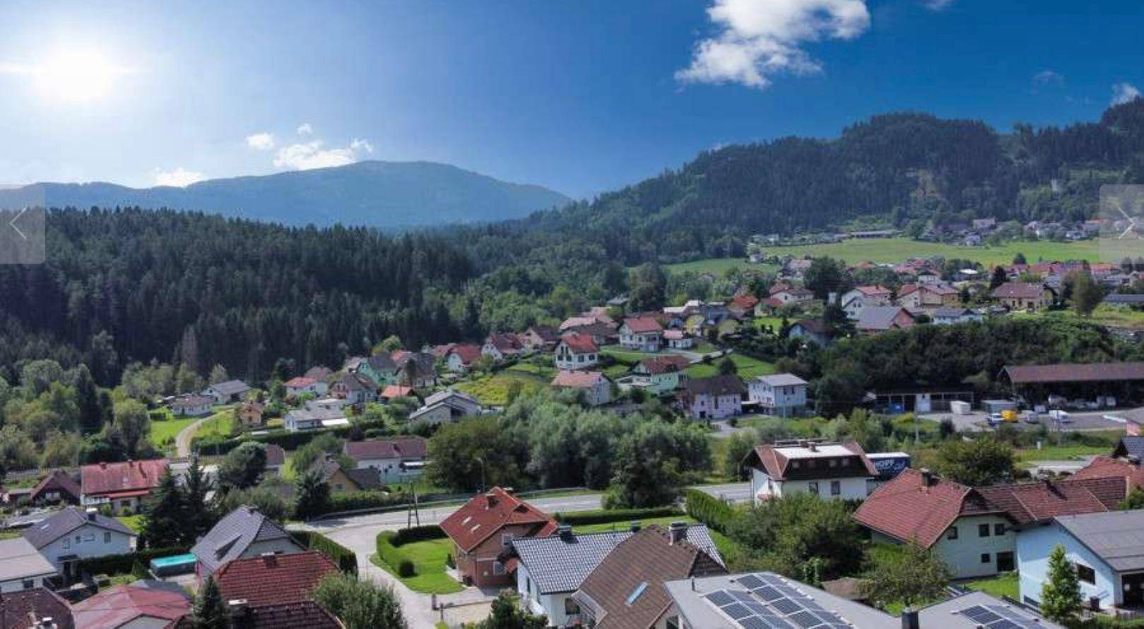 Panoramablick auf ein malerisches Dorf in einem Tal, umgeben von grünen Wäldern und Bergen.