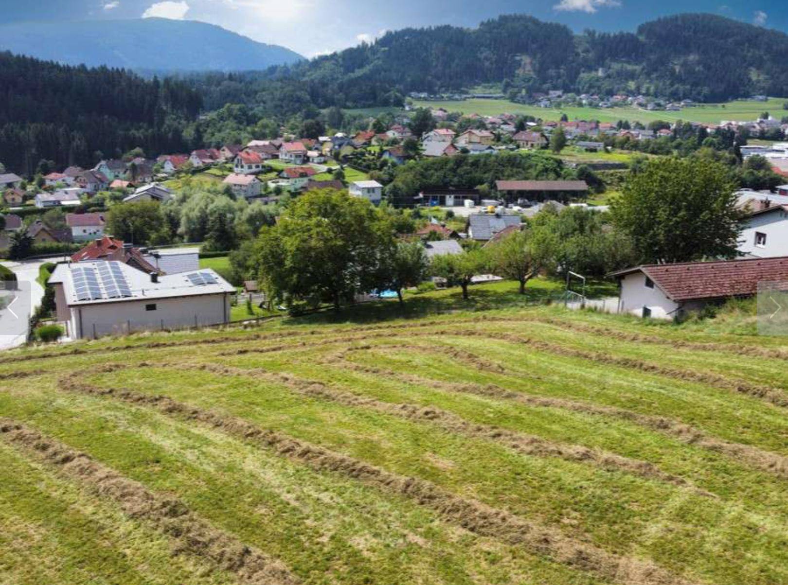 Blick über ein gemähtes Feld auf ein Dorf in einem grünen Tal mit bewaldeten Bergen im Hintergrund.