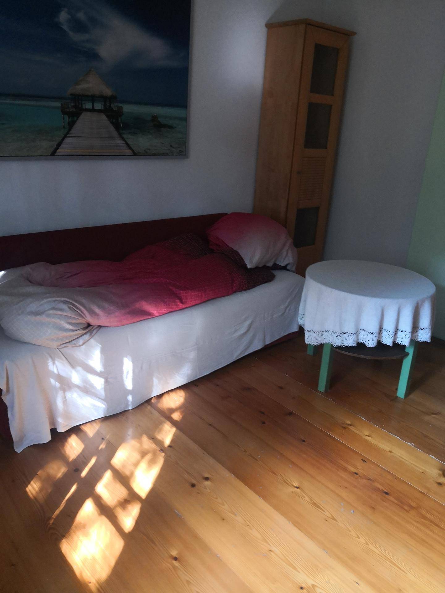Nahaufnahme eines Bettes mit roter Bettwäsche und kleinem Beistelltisch auf sonnigem Holzboden.