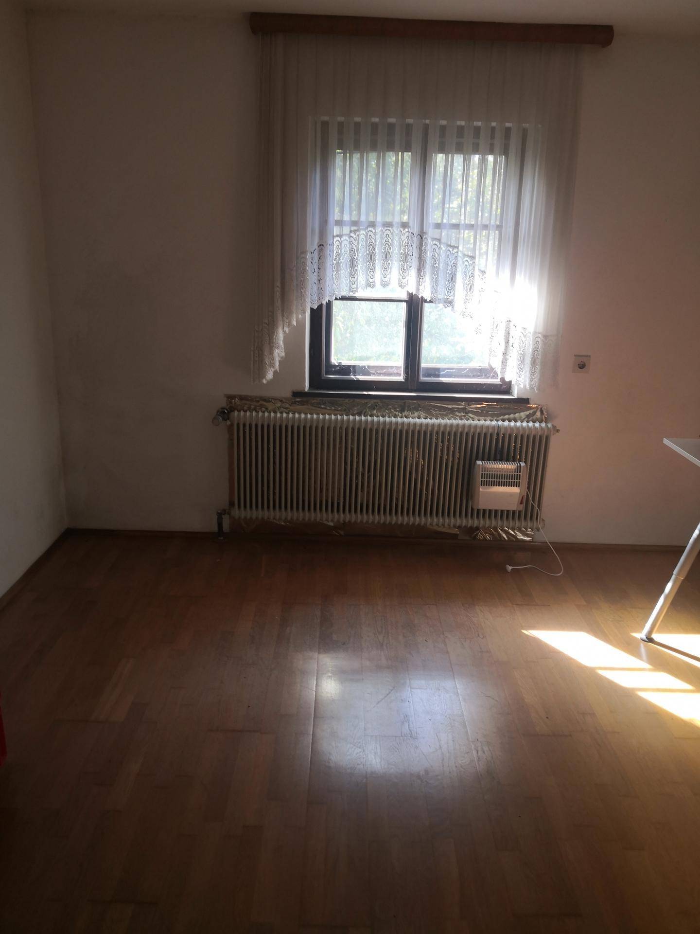 Leerer Raum mit Fenster, Heizkörper und Holzboden, durchflutet von natürlichem Sonnenlicht.
