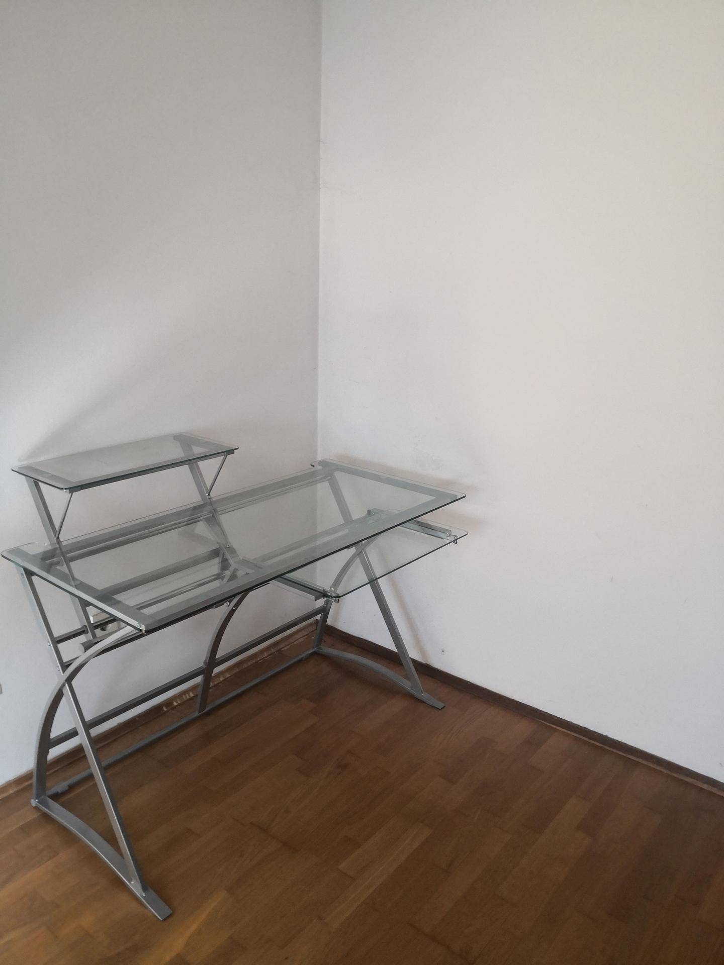 Ecke eines Raumes mit modernem Schreibtisch aus Glas und Metall auf Holzboden.
