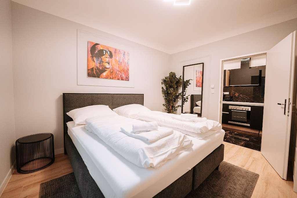 Schlafzimmer mit Doppelbett und Zugang zu einer modernen, dunklen Küchenzeile.