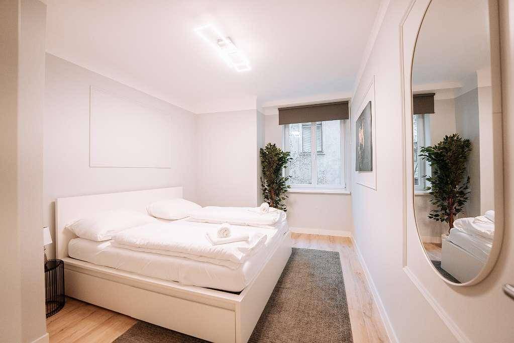 Helles Schlafzimmer mit weißem Doppelbett, Teppich und einem dekorativen Spiegel.