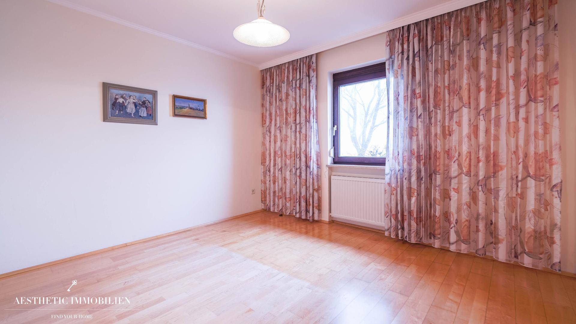 Helles Schlafzimmer mit Parkettboden, großem Fenster und floralen Vorhängen.