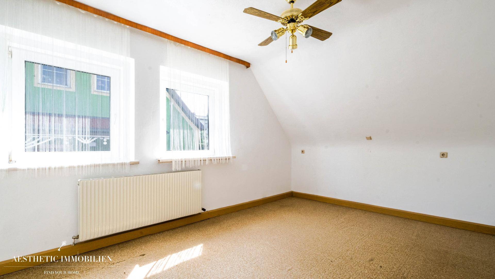 Heller Raum im Dachgeschoss mit Teppichboden, Deckenventilator und zwei Fenstern.