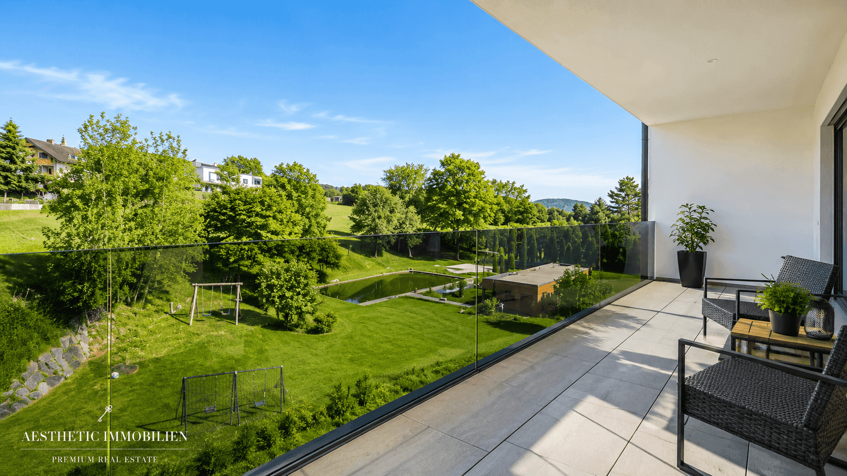 Geräumiger Balkon mit Glasgeländer und weitem Blick auf grüne Landschaft mit Teich und Spielplatz.