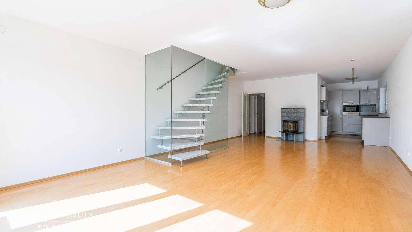 Leerer Wohnbereich mit Kamin, moderner Glastreppe und offenem Übergang zur Küche.