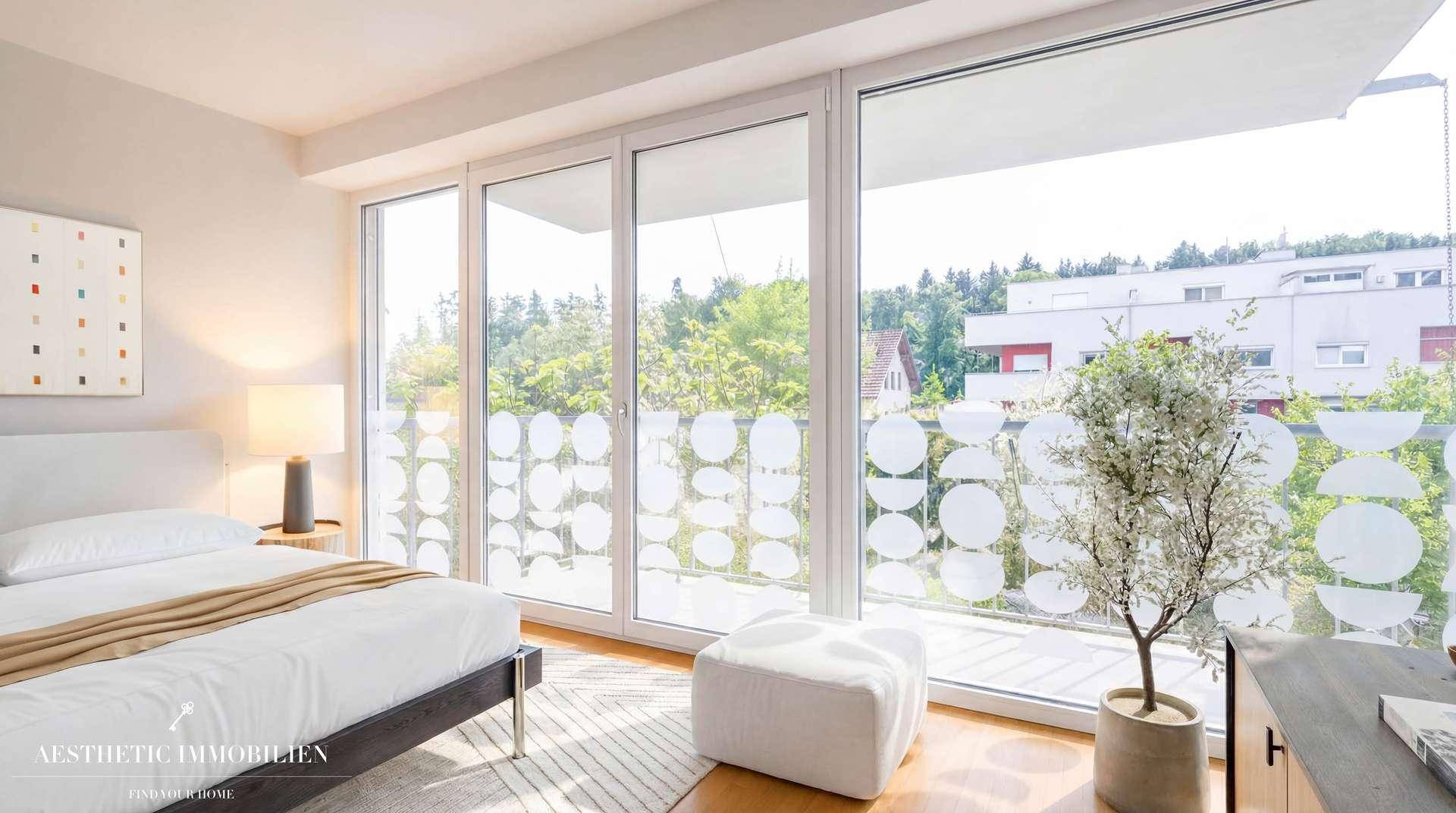 Helles Schlafzimmer mit großen Fenstern, die einen Blick auf Bäume und Gebäude bieten.