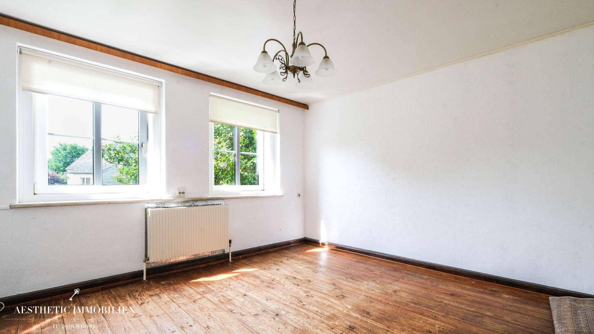 Geräumiges Wohnzimmer mit Holzdielenboden, zwei großen Fenstern und viel Tageslicht.