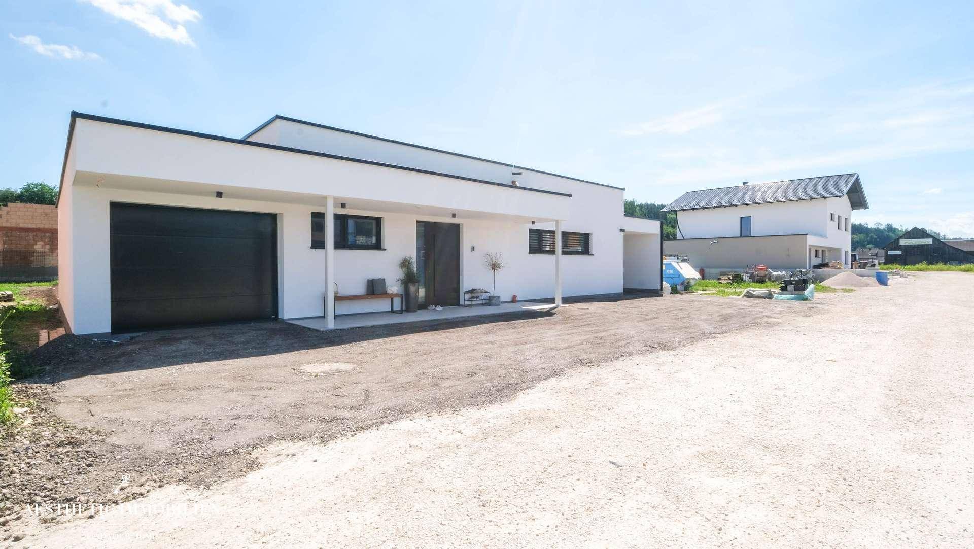 Moderne weiße Hausfassade mit Garage und Carport, umgeben von unbefestigtem Gelände.