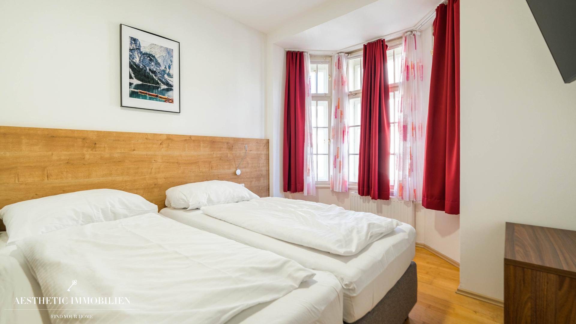 Gemütliches Schlafzimmer mit zwei Einzelbetten, Holzkopfteil und großen Fenstern mit roten Vorhängen.