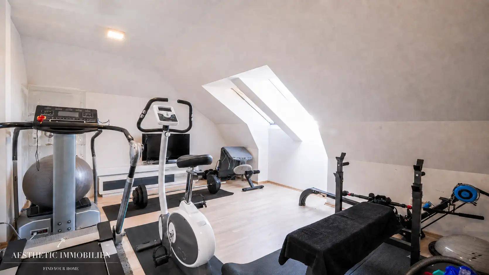 Fitnessraum im Dachgeschoss mit verschiedenen Trainingsgeräten und natürlichem Licht durch das Dachfenster.