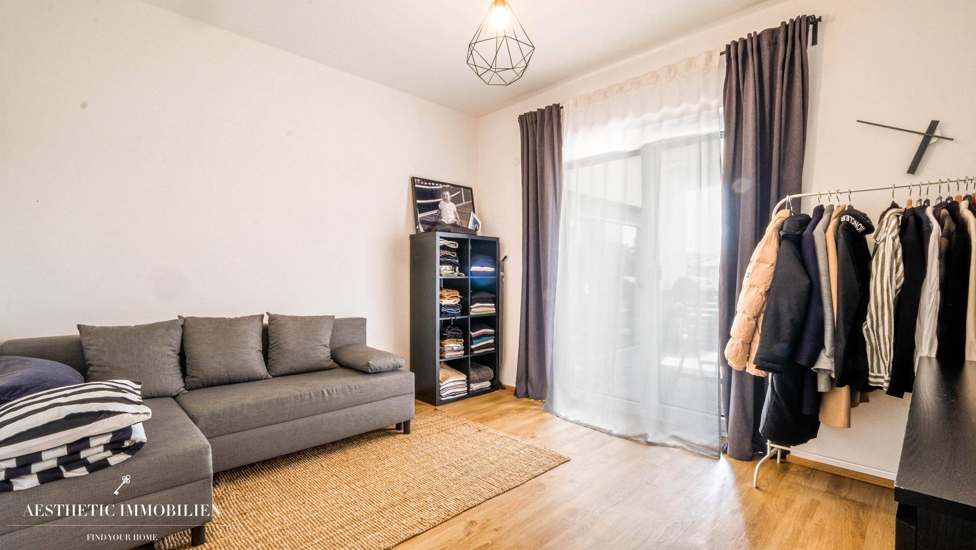 Helles Zimmer mit grauem Sofa, Kleiderstange und großem Fenster mit Vorhängen.