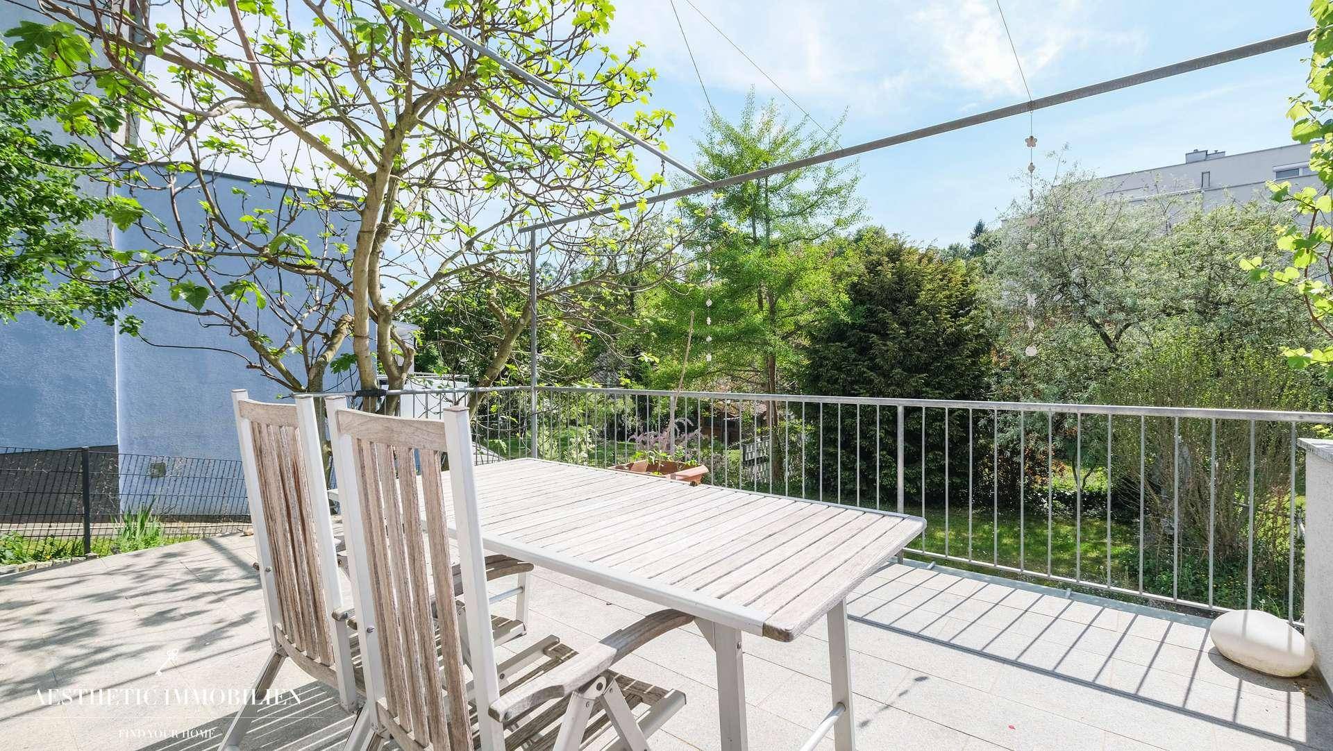 Sonniger Terrassenbereich mit Esstisch und Stühlen, umgeben von üppigem Grün und Bäumen.