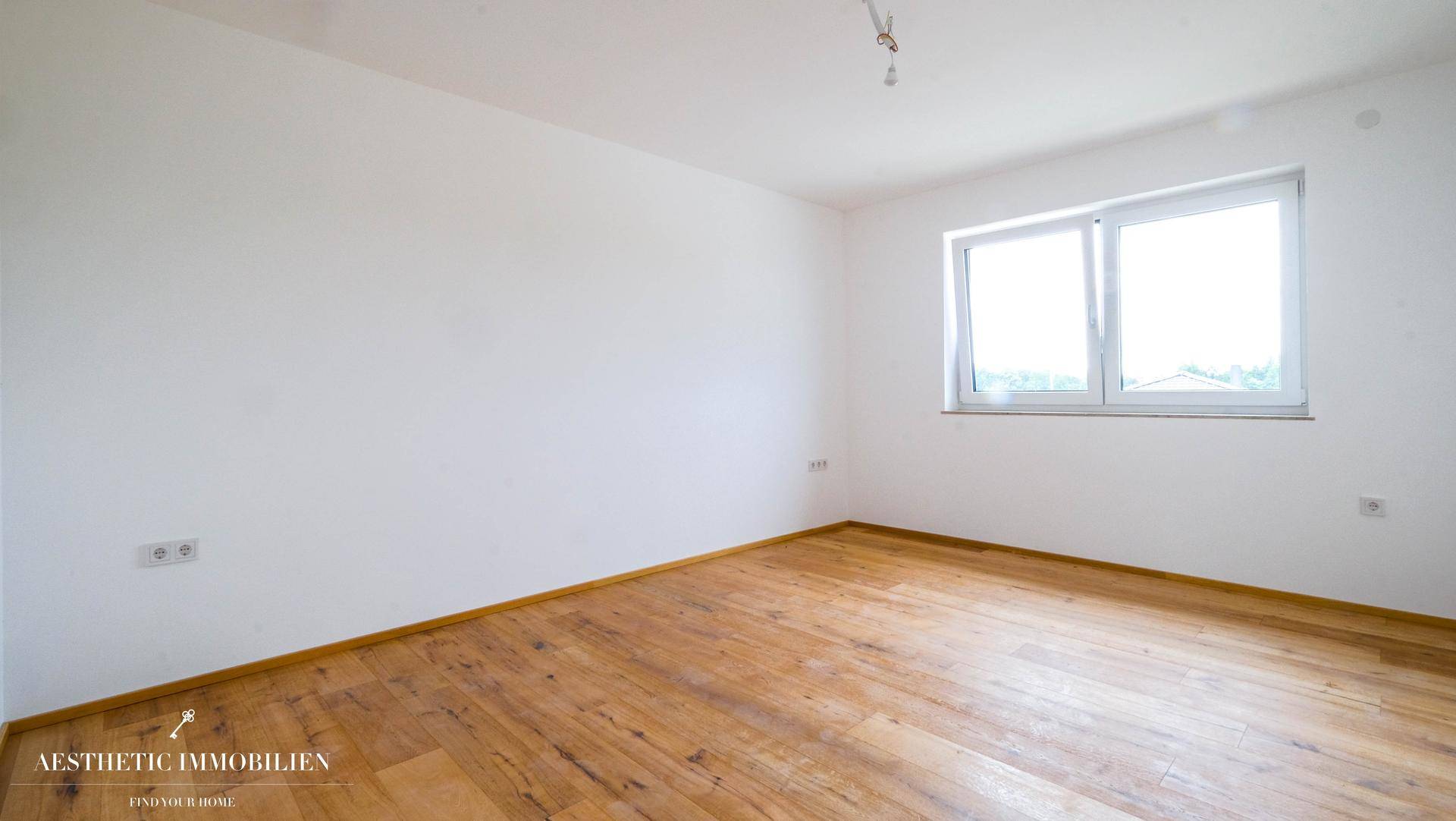 Leeres Zimmer mit Holzboden und einem Fenster, das viel Tageslicht in den Raum bringt.