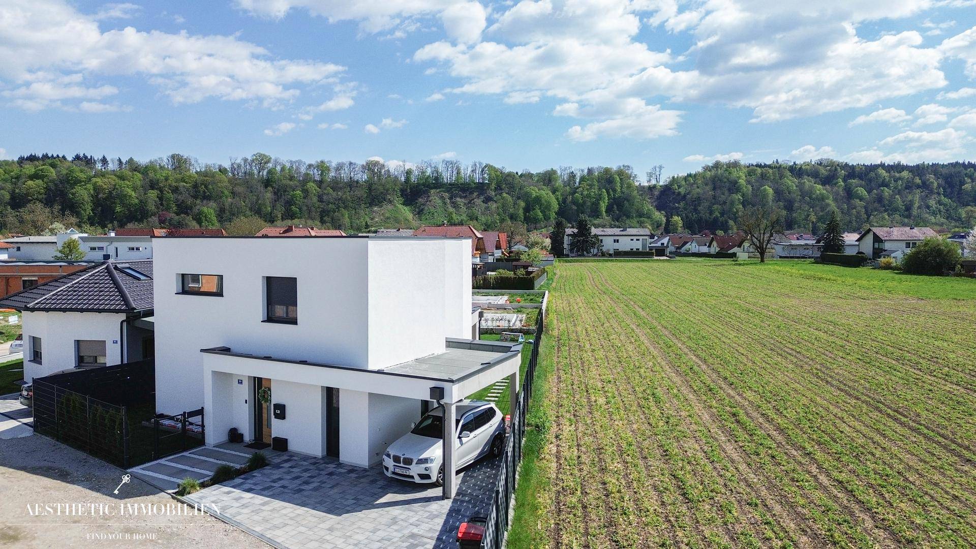 Modernes Einfamilienhaus mit Carport, umgeben von grünen Feldern und Natur.