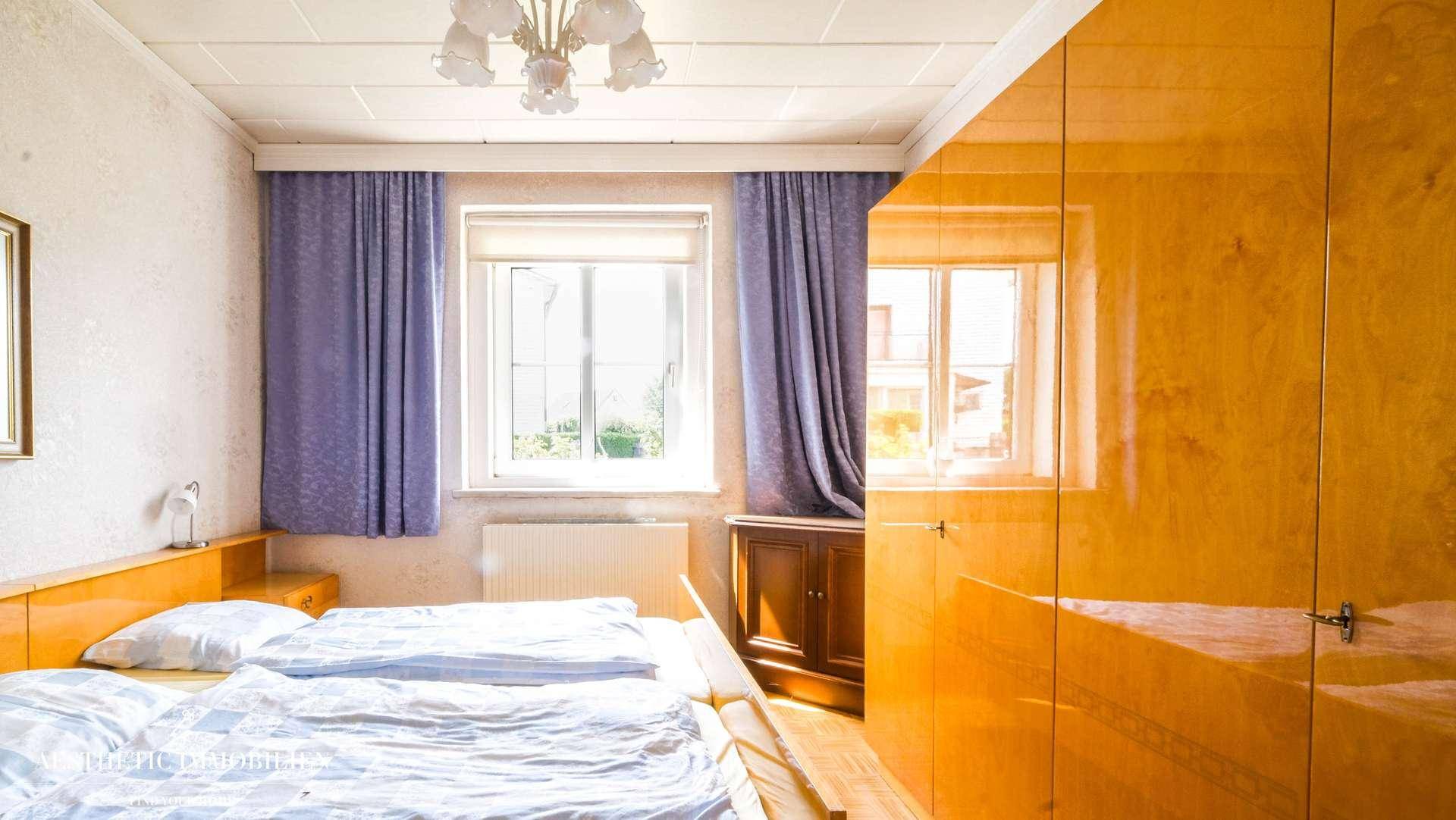 Helles Schlafzimmer mit Holzmöbeln, lila Vorhängen und Blick ins Grüne.